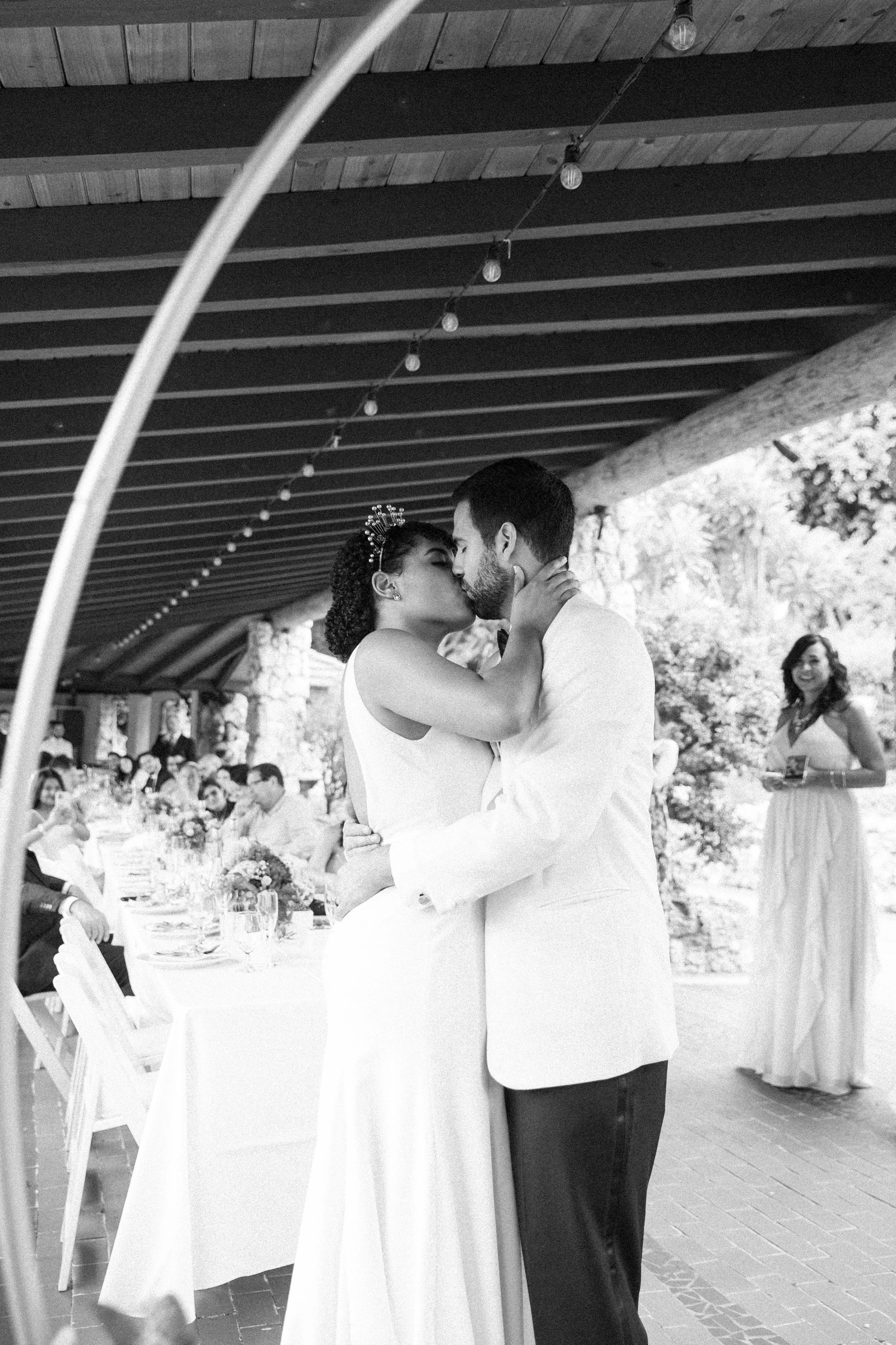 A bride and groom kiss during their wedding reception, with guests seated at a decorated table in the background, and a bridesmaid standing nearby with a glass in hand.