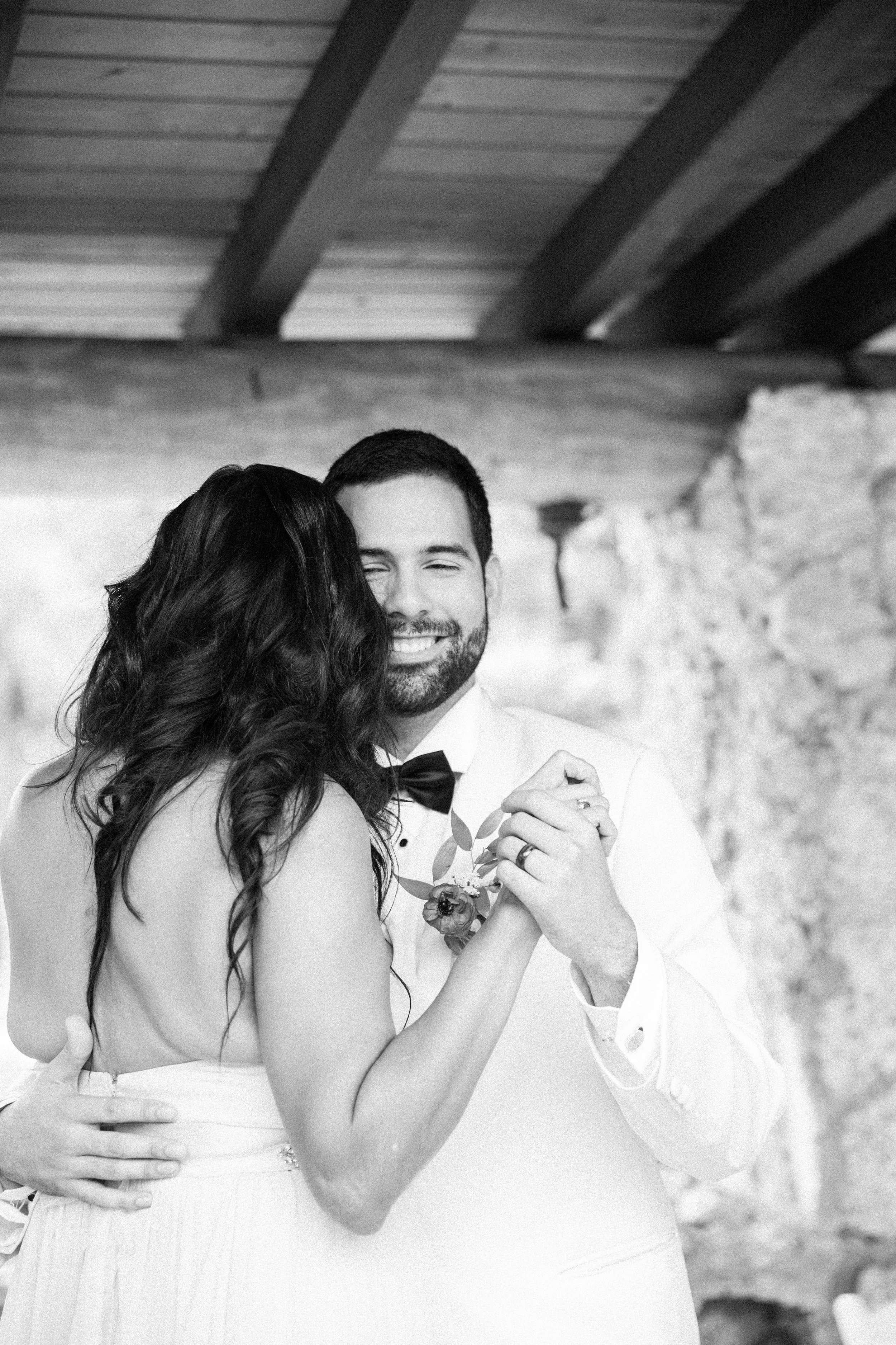 A black and white photo of a couple dancing closely, smiling, with the woman leaning her head against the man's face, in an indoor setting with stone walls and a wooden ceiling.