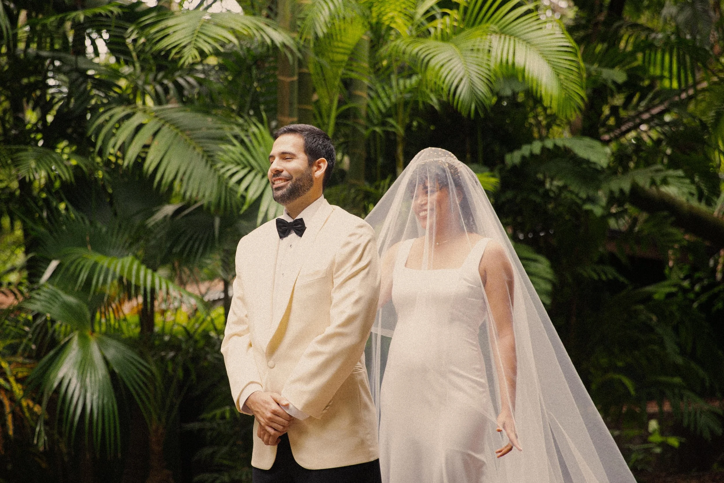 A bride and groom standing in a lush, green jungle setting, wearing wedding attire. The groom is smiling in a cream tuxedo with a black bow tie, and the bride, standing behind him, is smiling with a sheer veil over her face, dressed in a white weddin