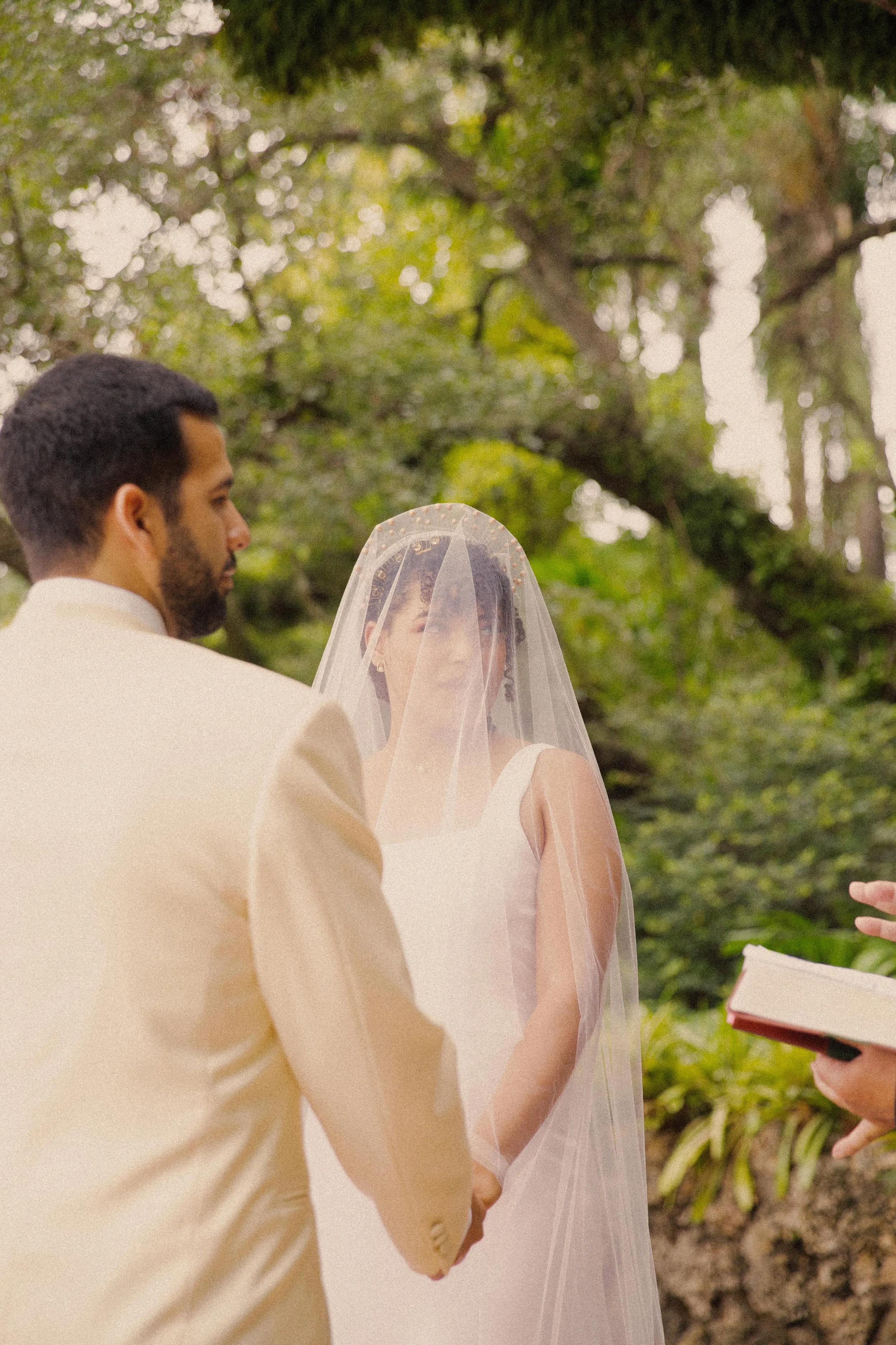 A wedding ceremony outdoors with a bride and groom holding hands, officiant reading, surrounded by lush green trees.