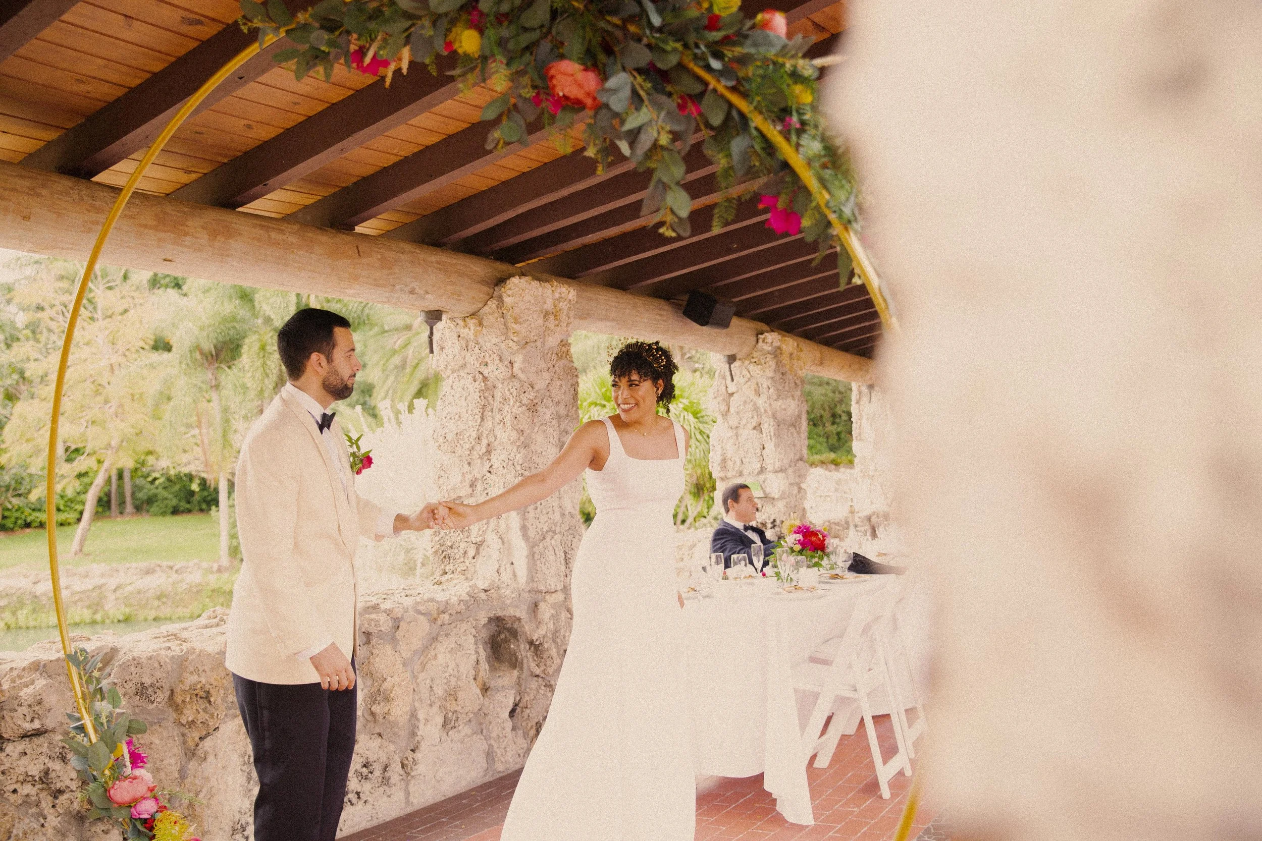 A bride and groom holding hands during their wedding ceremony with a seated man in the background at a decorated outdoor venue.