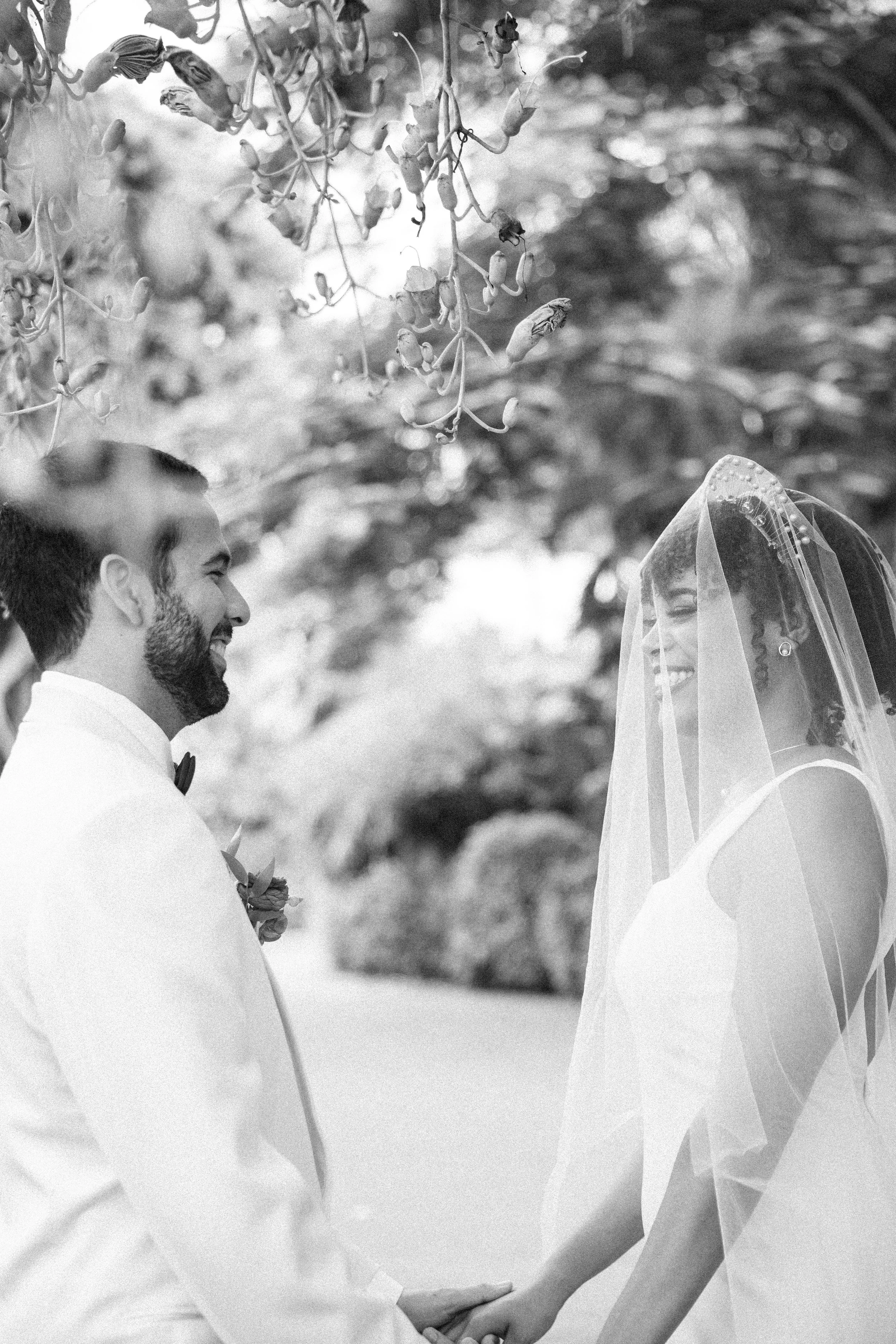 A black and white photo of a wedding, showing a couple holding hands and smiling at each other beneath a tree with hanging flowers. The groom is on the left with a beard, wearing a white suit and bow tie, while the bride is on the right with a veil a