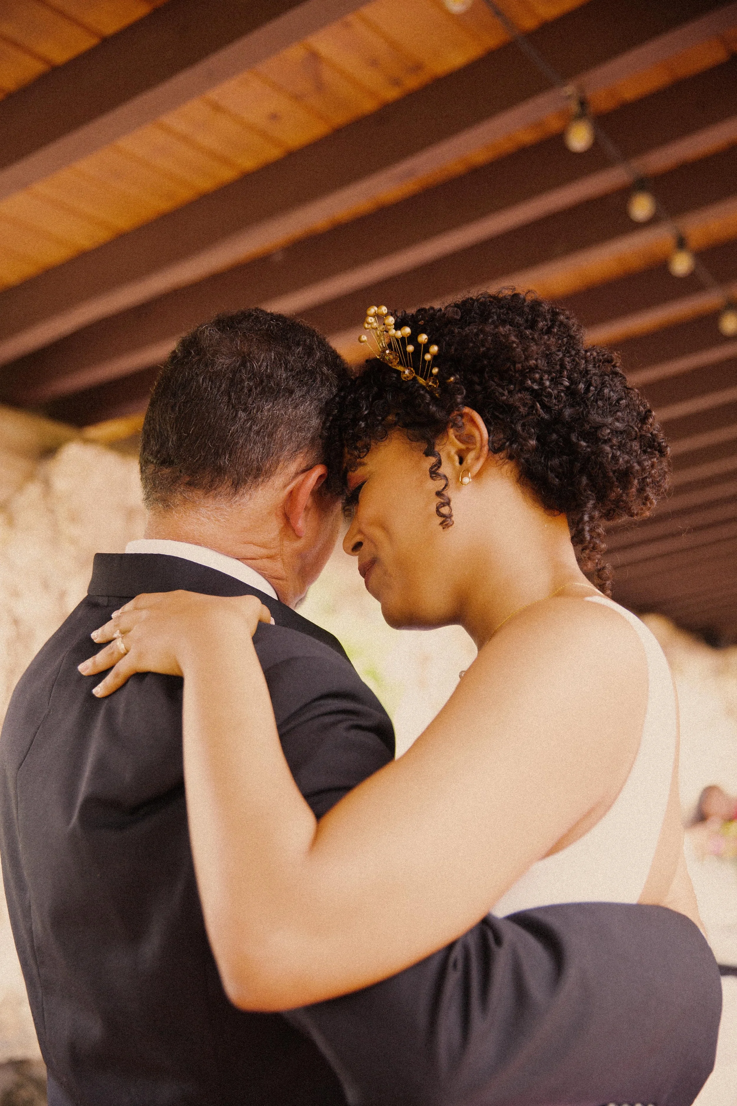 Bride and groom sharing a dance with foreheads touching, wedding reception setting with wooden ceiling and hanging lights.