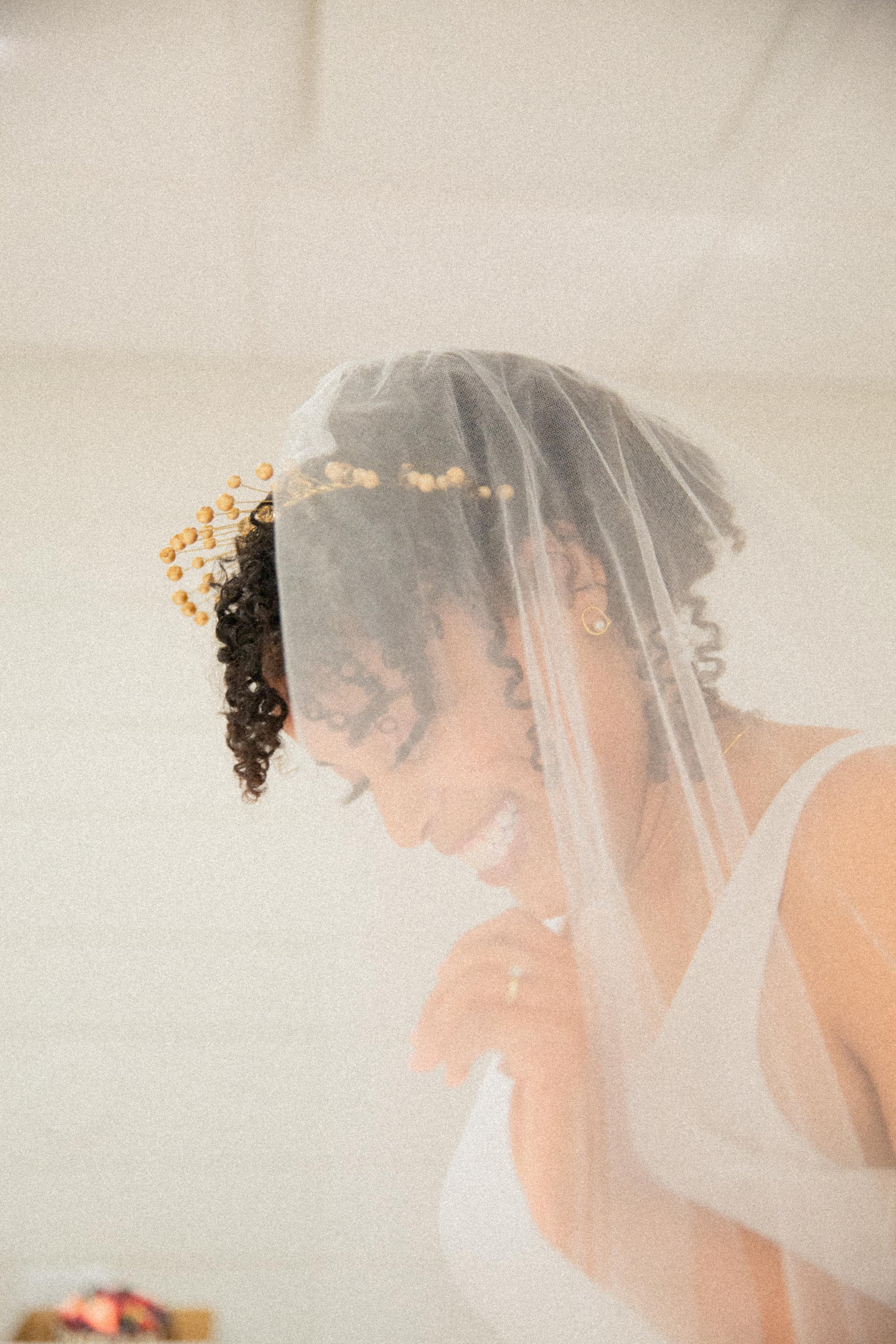 A woman with curly hair wearing a wedding veil and a white dress, smiling and looking down.