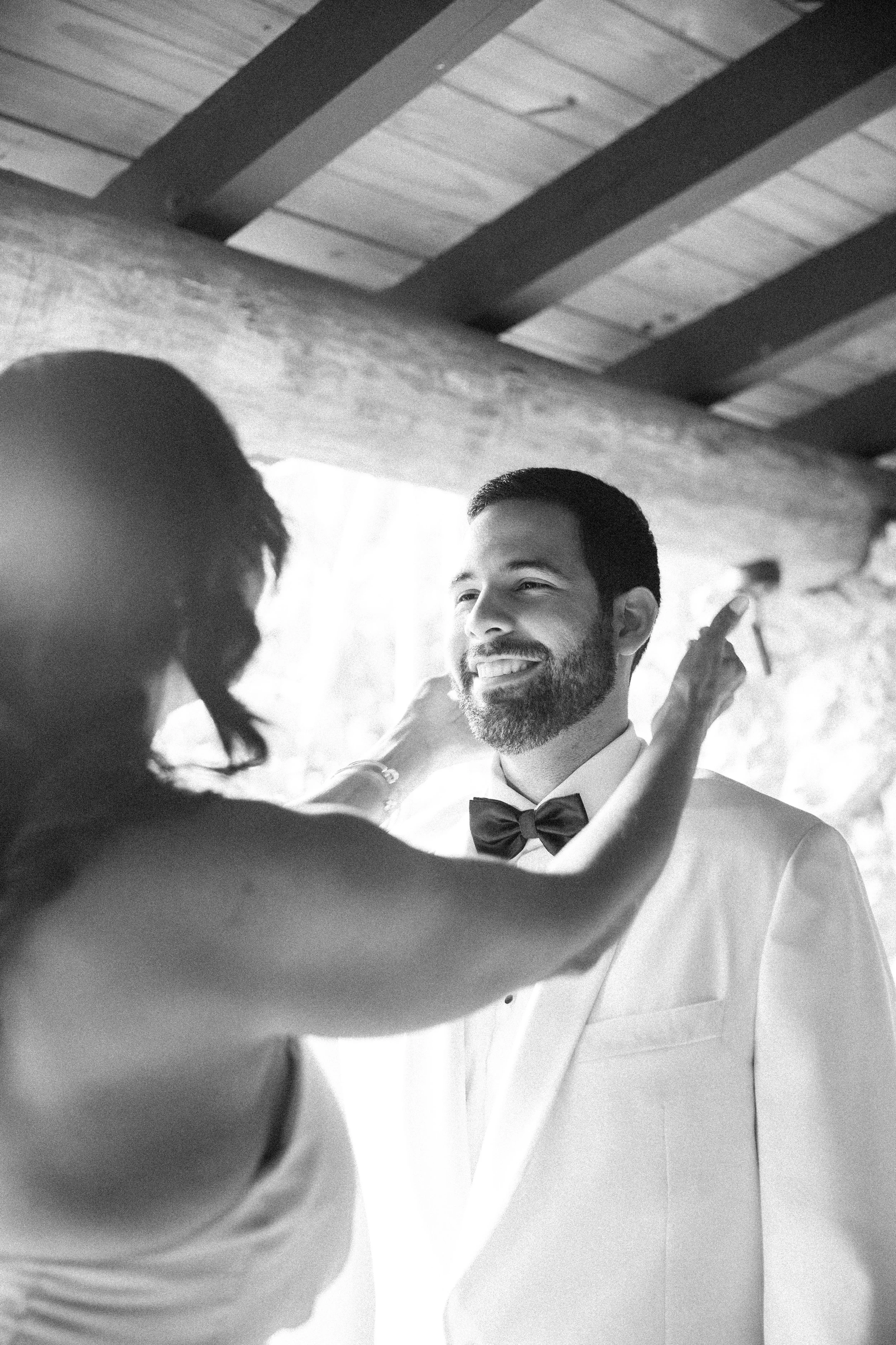 A black-and-white photo of a groom in a tuxedo with a bow tie, smiling as a woman with dark hair adjusts his collar.
