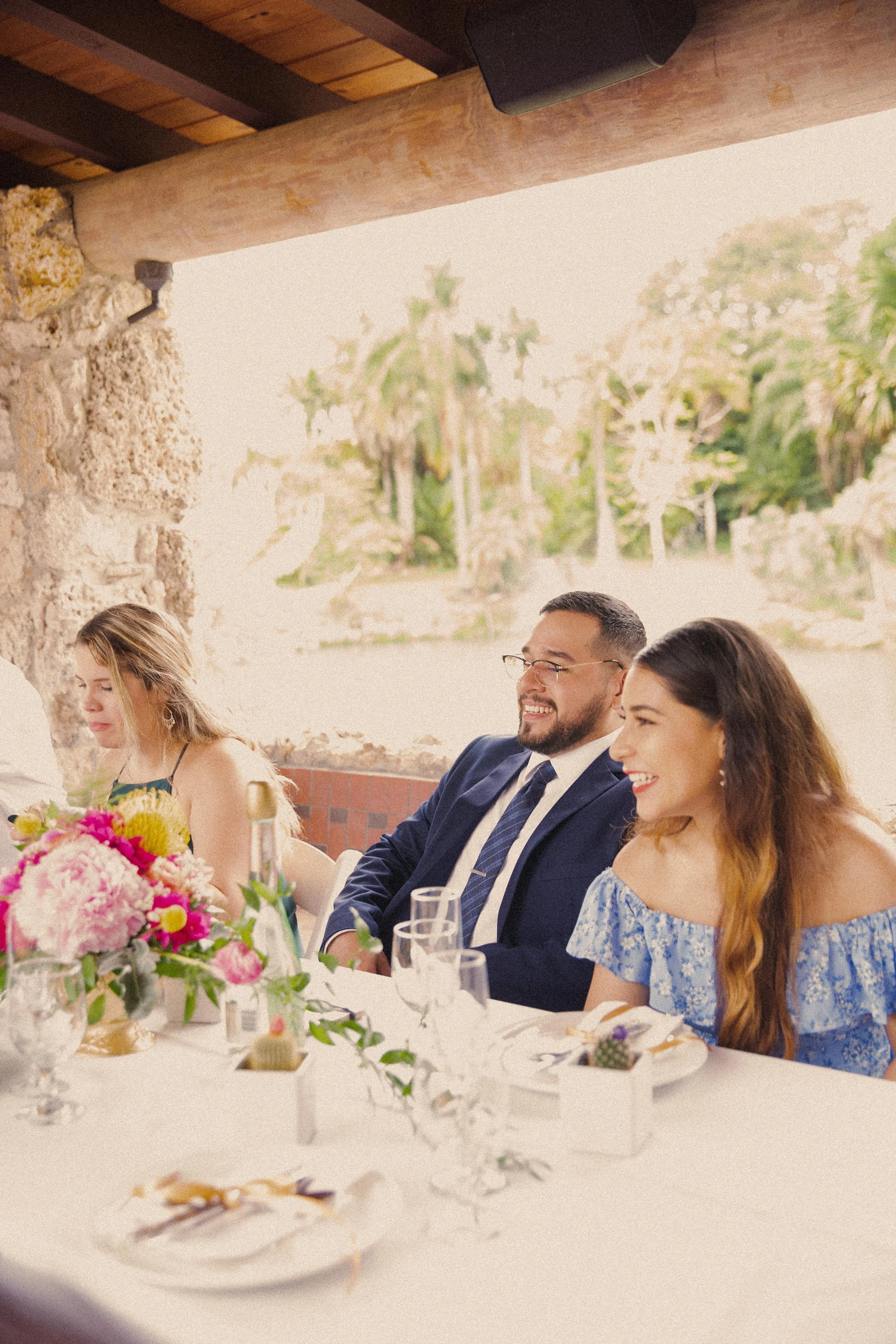 People sitting at a decorated table during a celebration, with two women and a man smiling, in front of a bright outdoor view with trees and stone wall.
