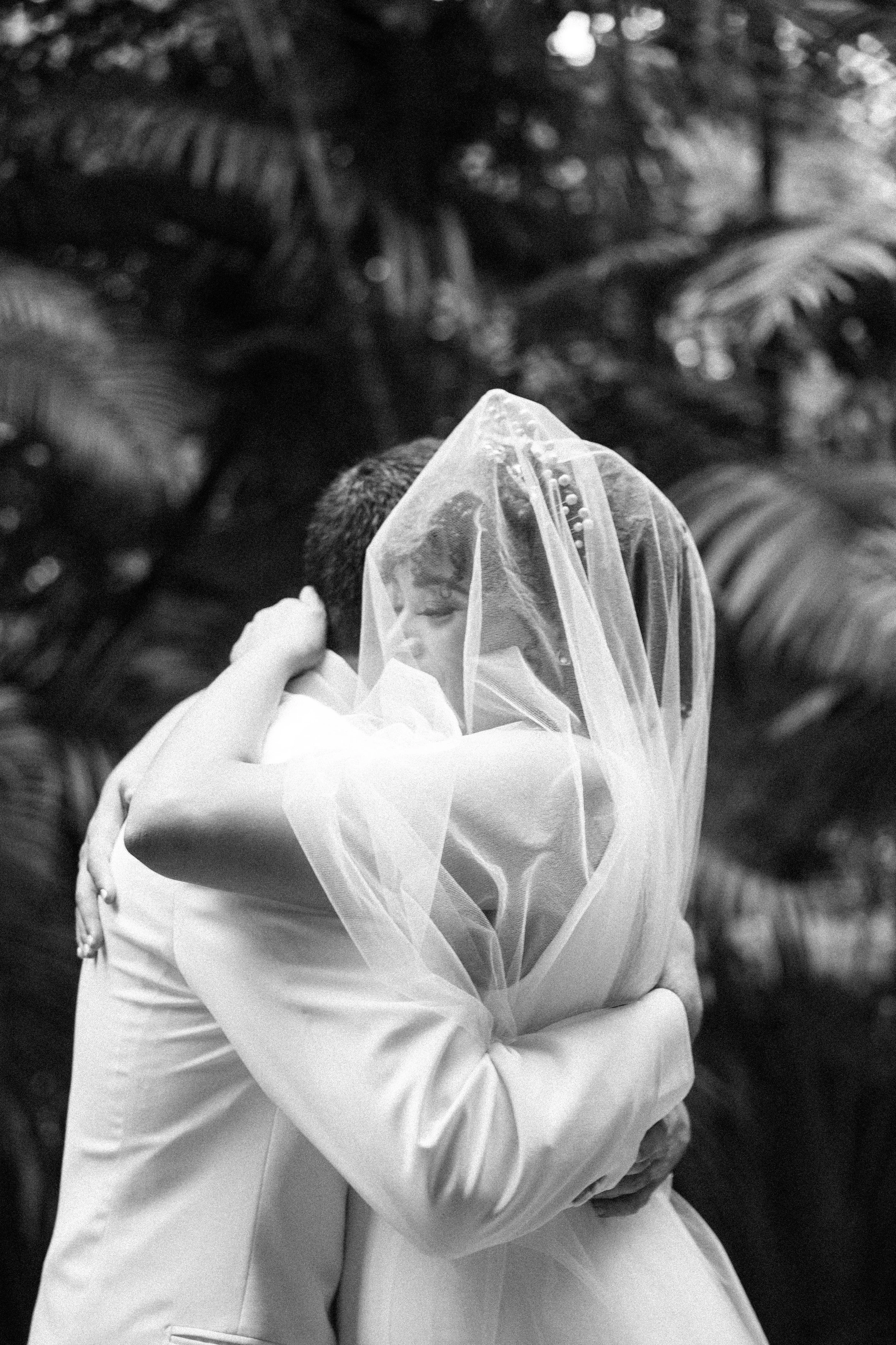 A couple hugging closely, with the woman wearing a veil, indicating a wedding or romantic moment, in an outdoor setting with blurred foliage in the background.