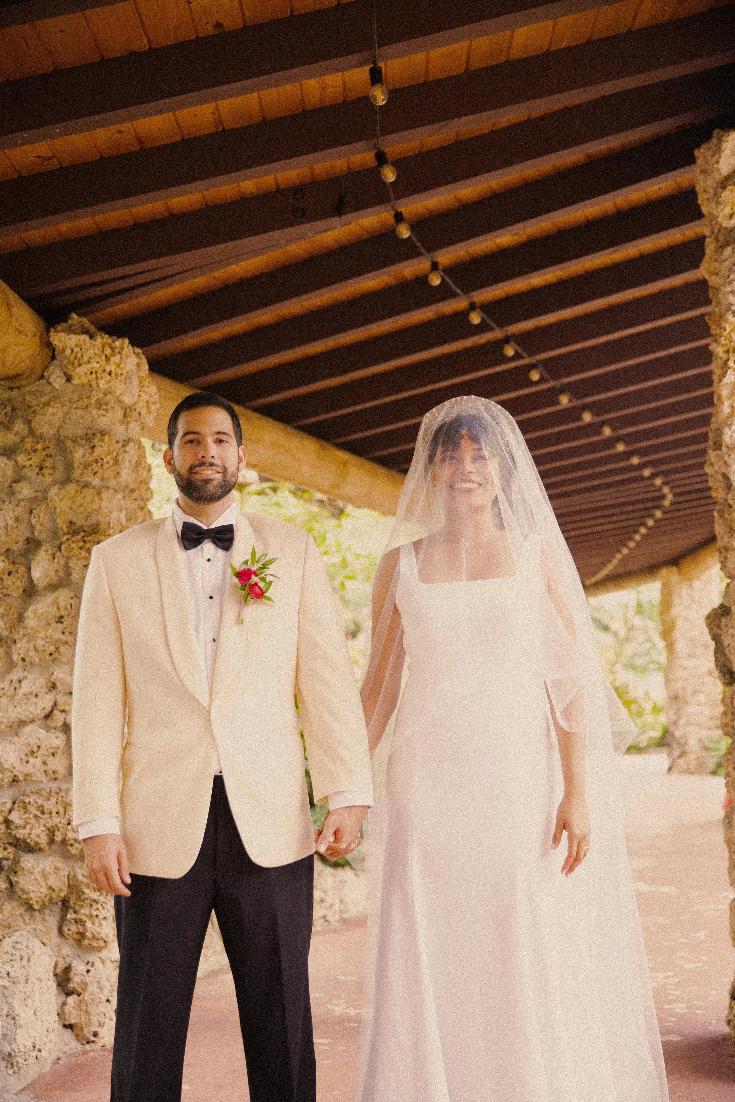 A bride and groom holding hands and standing together on their wedding day, under a rustic wooden roof with stone walls in the background. The groom is wearing a cream tuxedo with a black bow tie and a red boutonniere. The bride is wearing a sleevele