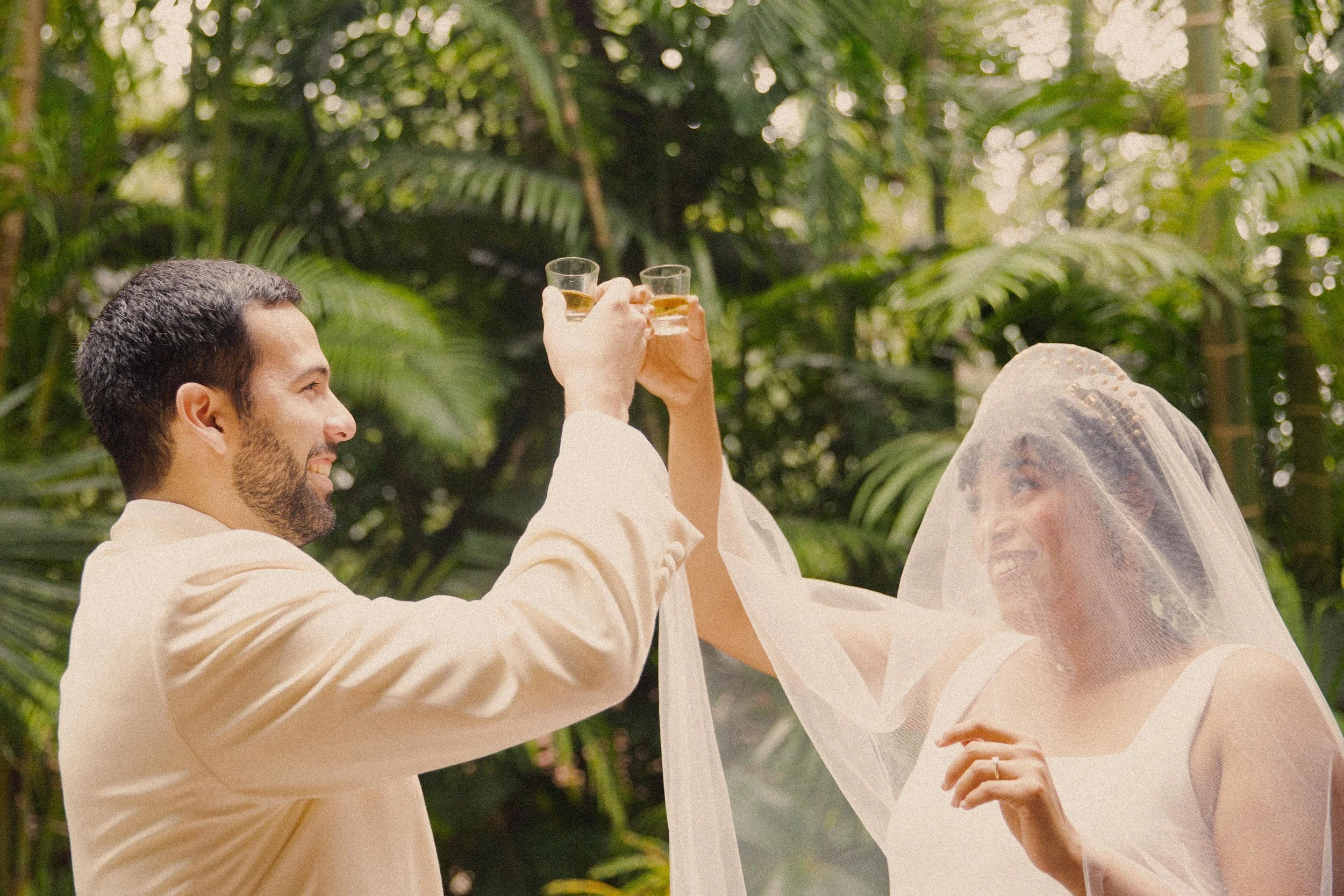 A couple toasting with drinks in a lush, green, outdoor setting, with the bride wearing a veil and smiling.