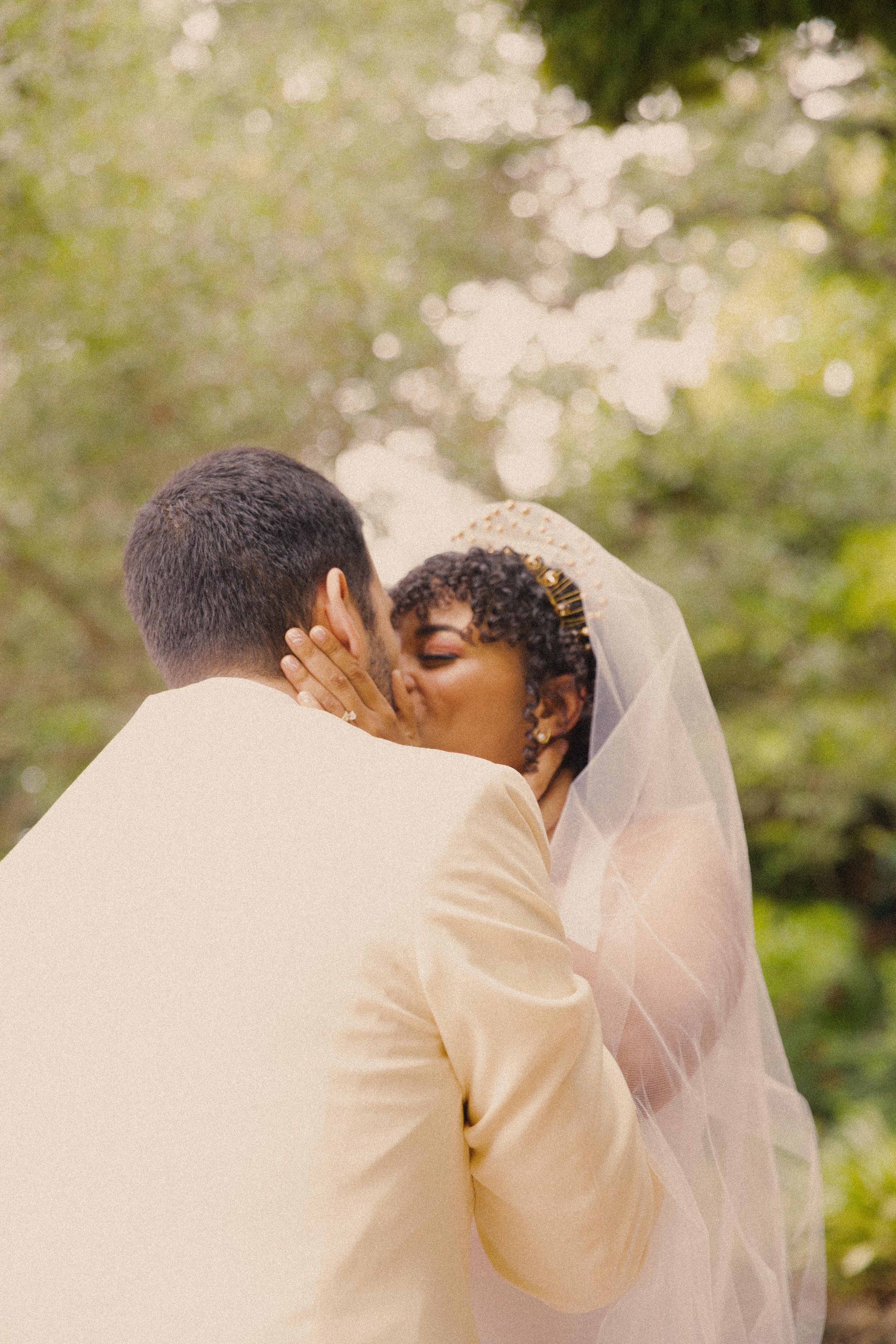 A couple sharing a kiss during their wedding ceremony outdoors, with greenery and blurred sunlight in the background.