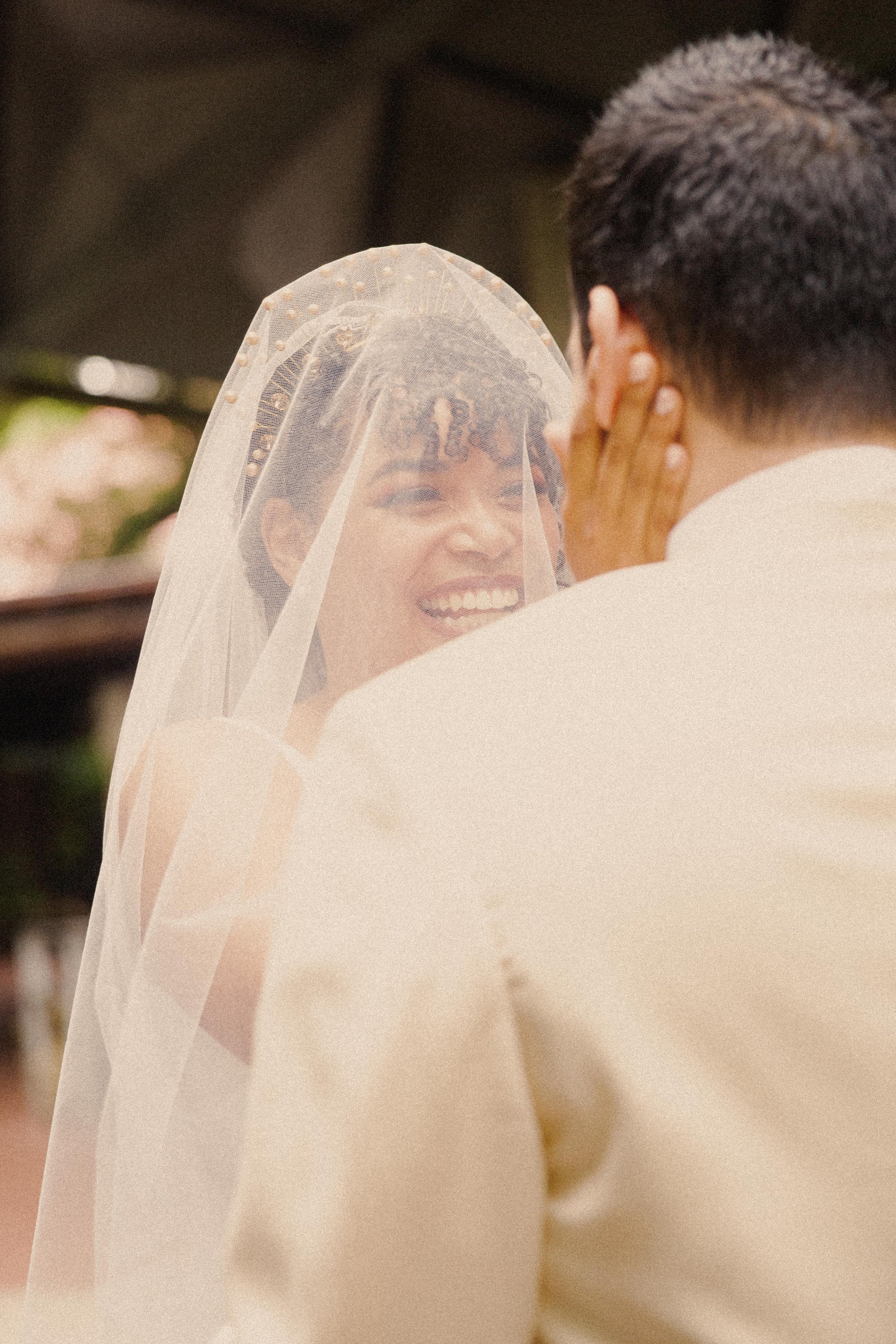 A bride smiling as she looks at her partner, who is touching her face, during a wedding ceremony. The bride is wearing a veil and bridal headpiece and has short, curly hair.