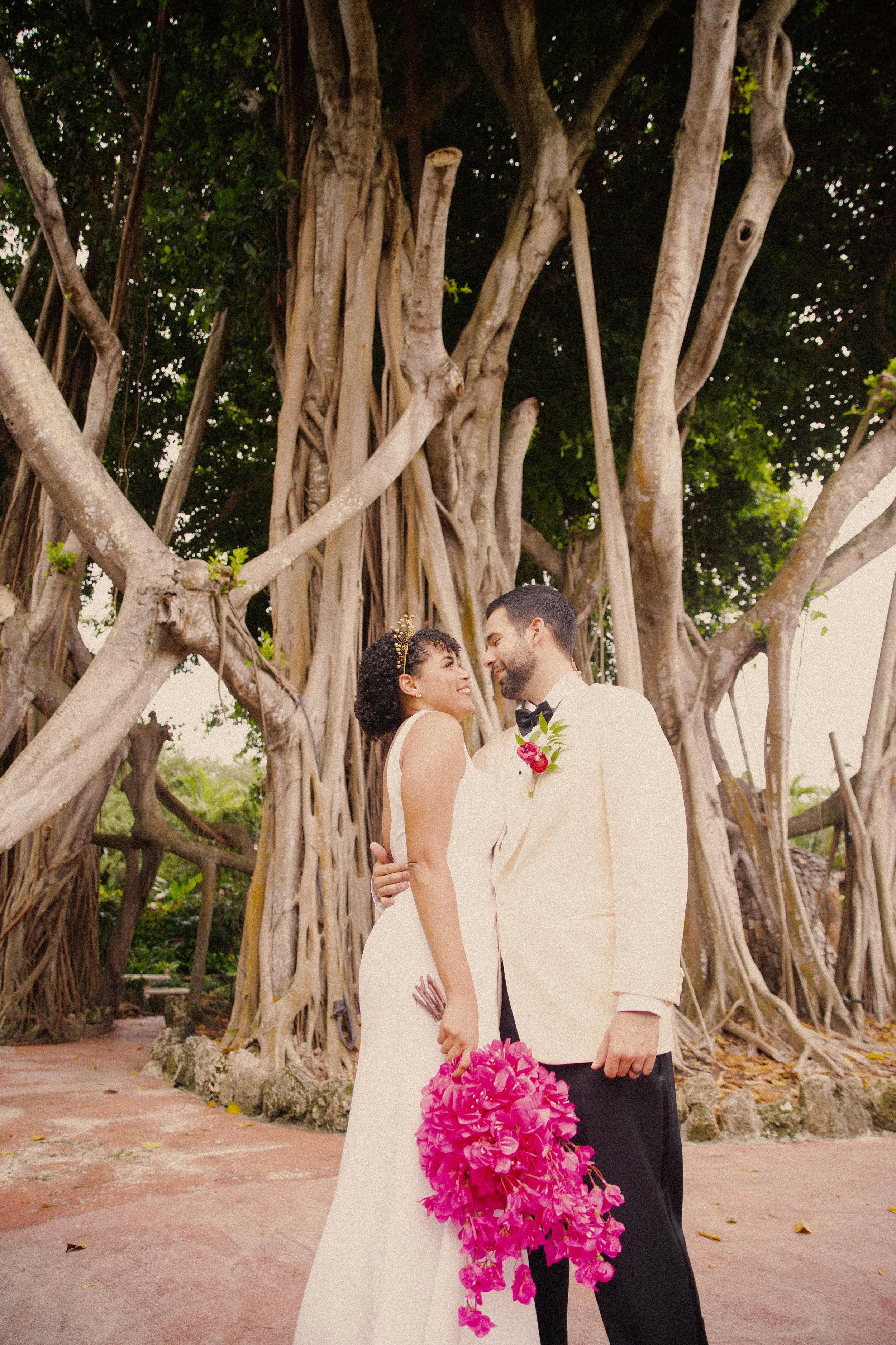 A bride and groom standing close together outdoors in front of large, sprawling trees, sharing a romantic moment. The bride is holding a large pink bouquet and wearing a white wedding gown with a tiara, while the groom is dressed in a light-colored t