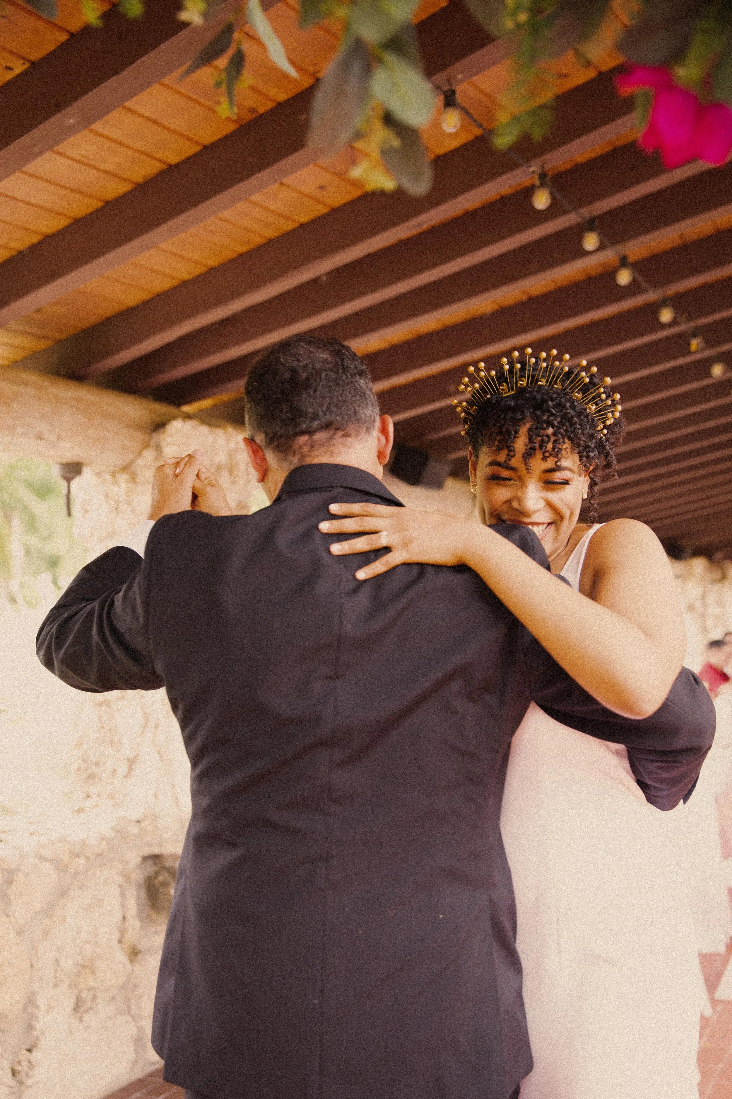 A woman in a white dress and a golden headpiece is dancing with a man in a black suit indoors, under a wooden ceiling with string lights and greenery hanging from above.