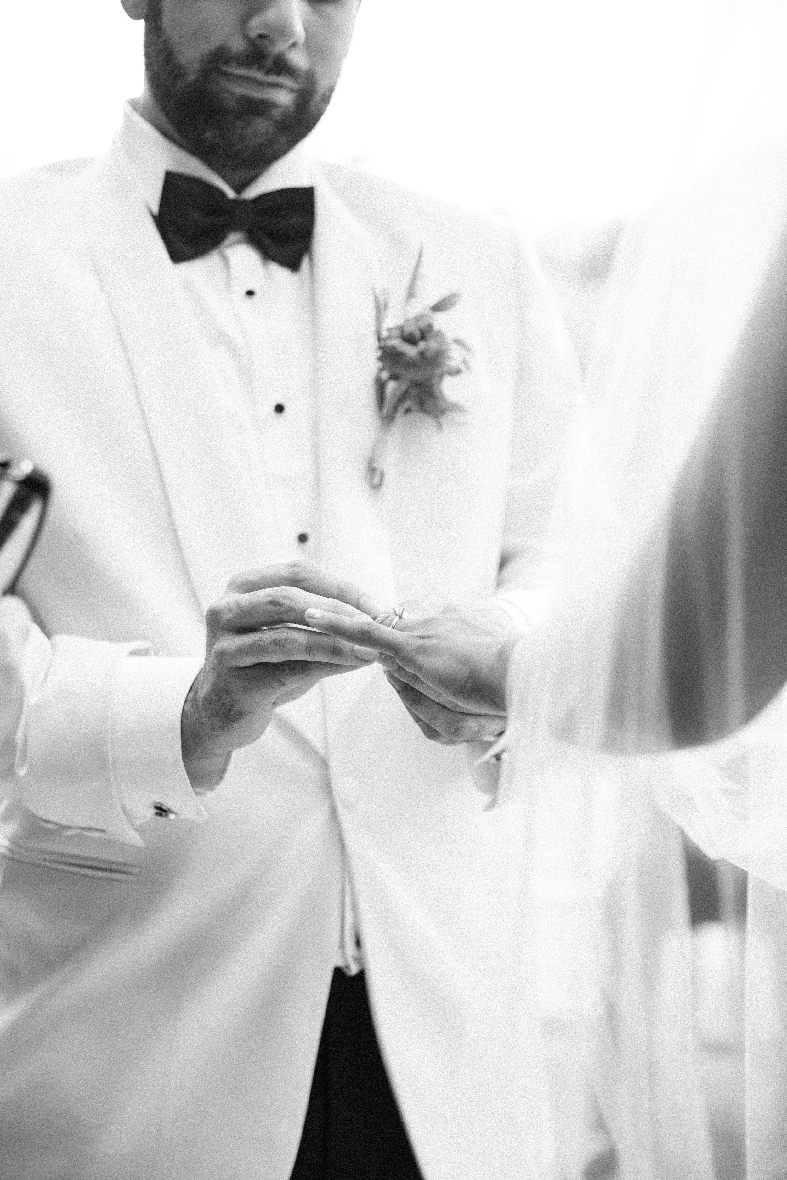 A man in a white tuxedo with a black bow tie is placing a ring on a woman's finger during a wedding ceremony. The man has a boutonniere on his lapel and a beard.