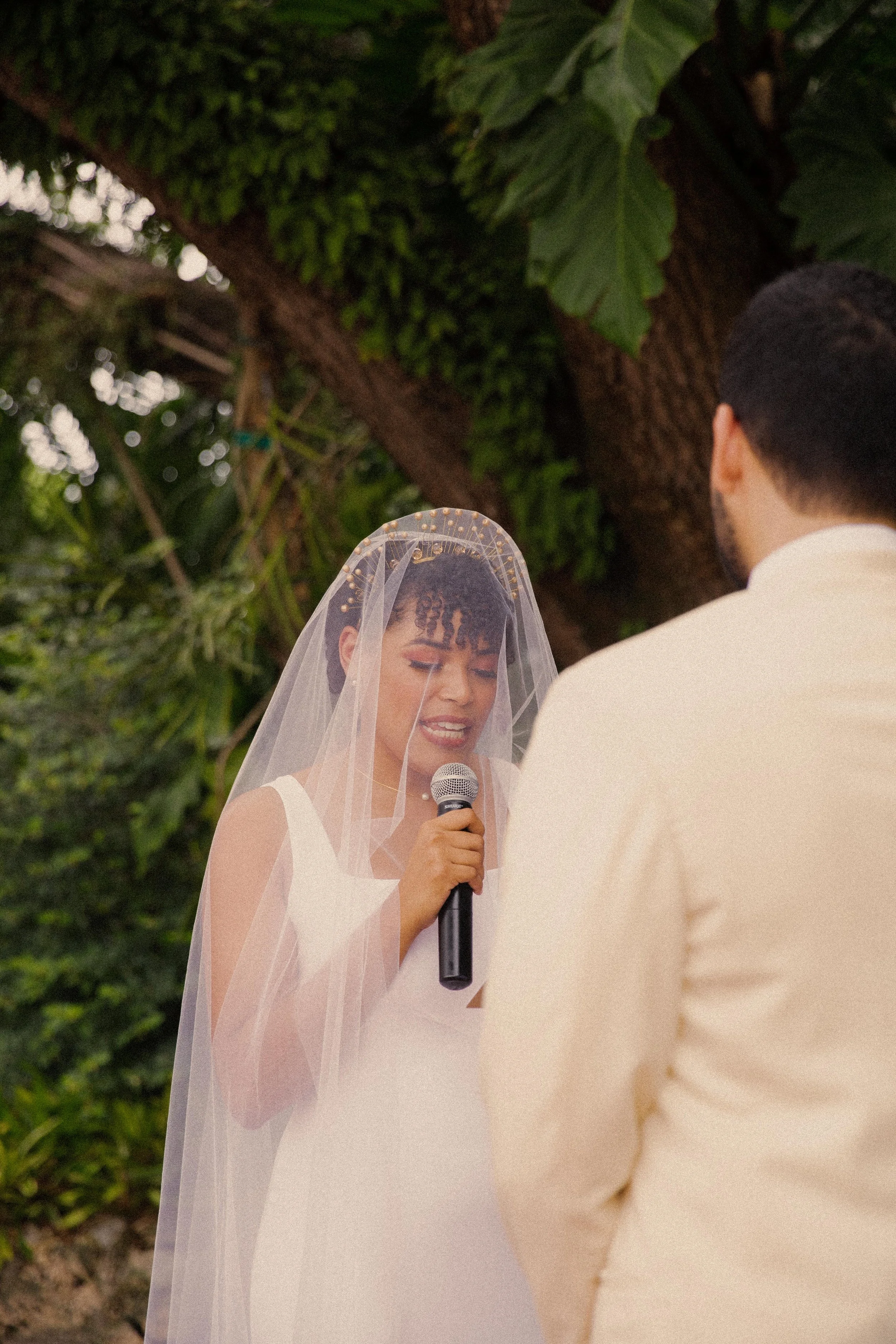A bride with a veil and tiara holding a microphone, standing outdoors in front of a large tree, during a wedding ceremony with an officiant or groom partially visible.