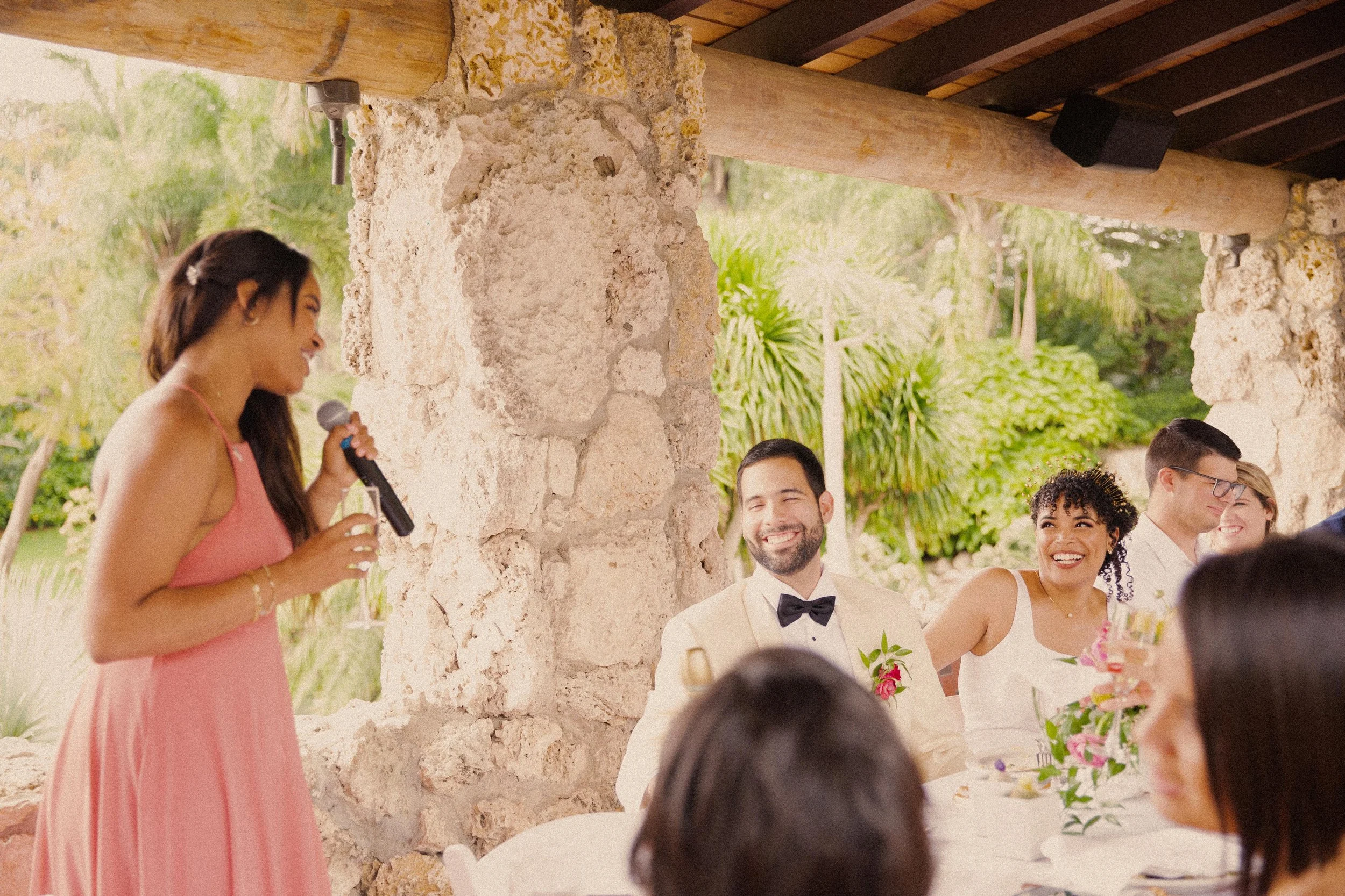 A woman in a pink dress giving a speech with a microphone at a wedding reception, with guests seated at tables and smiling.