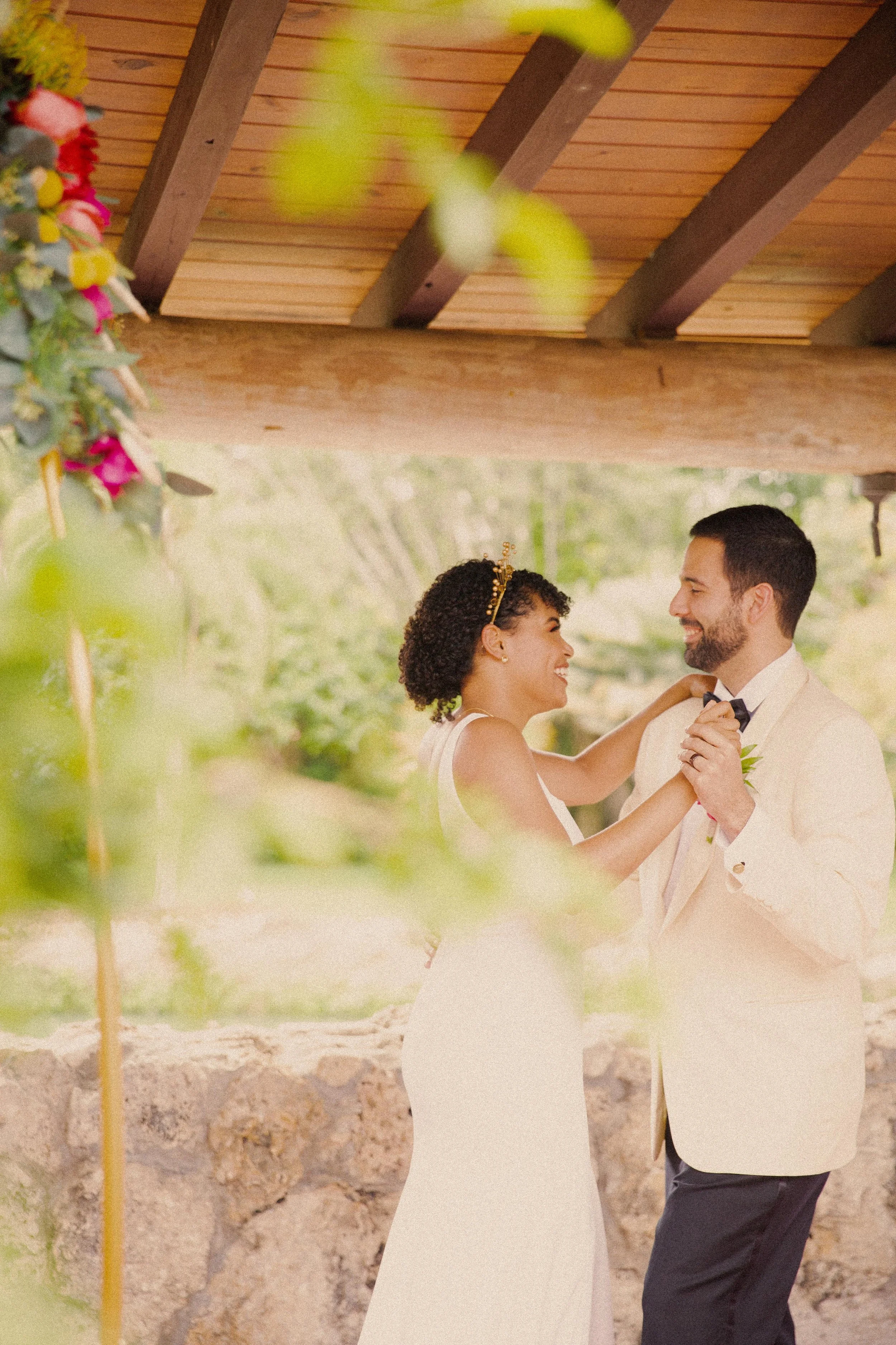 A bride and groom dancing under a wooden pavilion with greenery in the background.