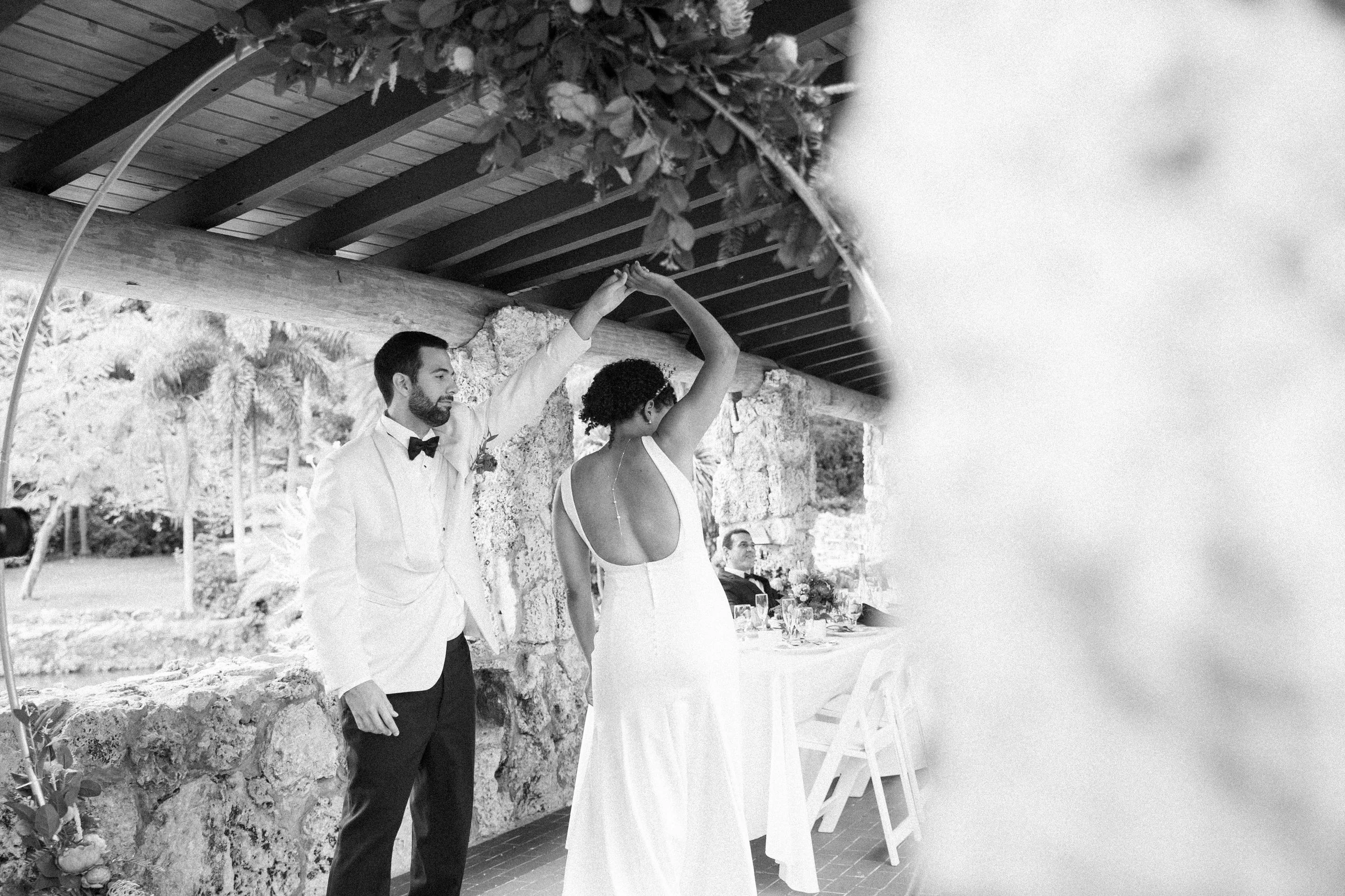 A black and white photo of a couple dancing at a wedding reception. The man is wearing a white tuxedo jacket with a bow tie and the woman is in a backless white dress. They are under a wooden roof with stone walls, with a table and a man visible in t