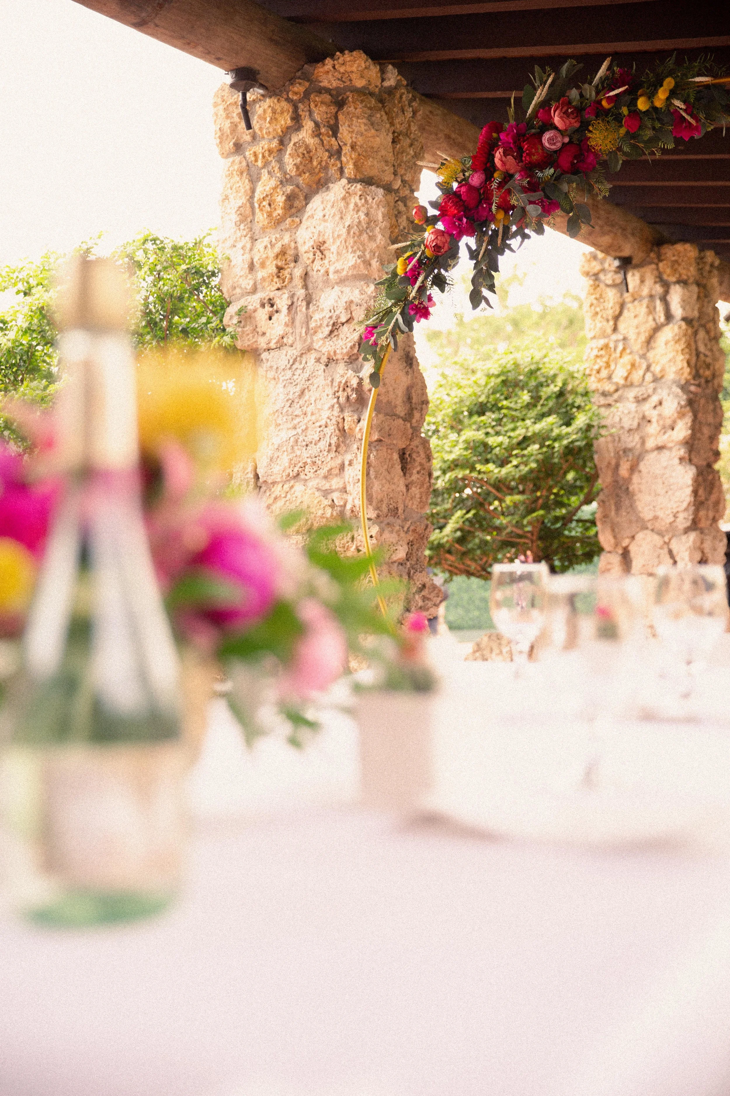 A decorated outdoor event space with stone pillars, hanging floral arrangement, and tables with vases of flowers and glasses, set in a natural, lush environment.