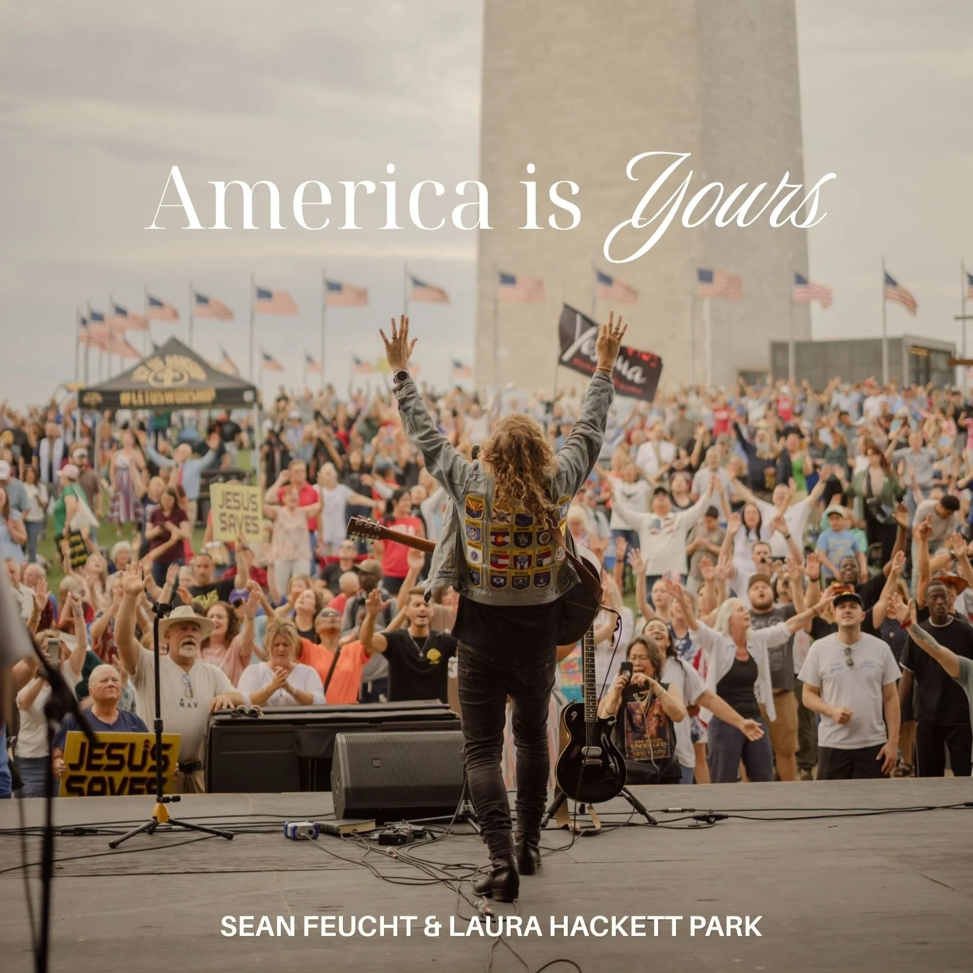 A musician with long, curly hair plays an electric guitar on an outdoor stage with a large crowd in front. The scene is at Sean Feucht and Laura Hackett Park event, with American flags and overcast sky in the background.