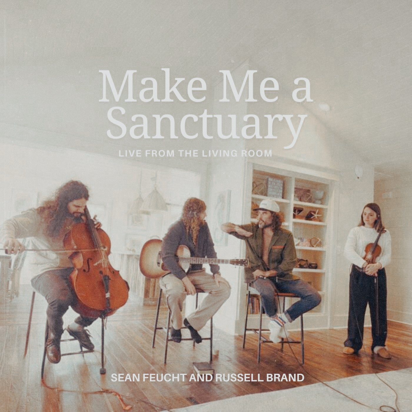 A band of four musicians performing in a cozy living room with wooden floors. The man on the left is playing a cello, the woman in the middle is seated with an acoustic guitar, the man next to her is talking and wearing a hat, and the woman on the ri