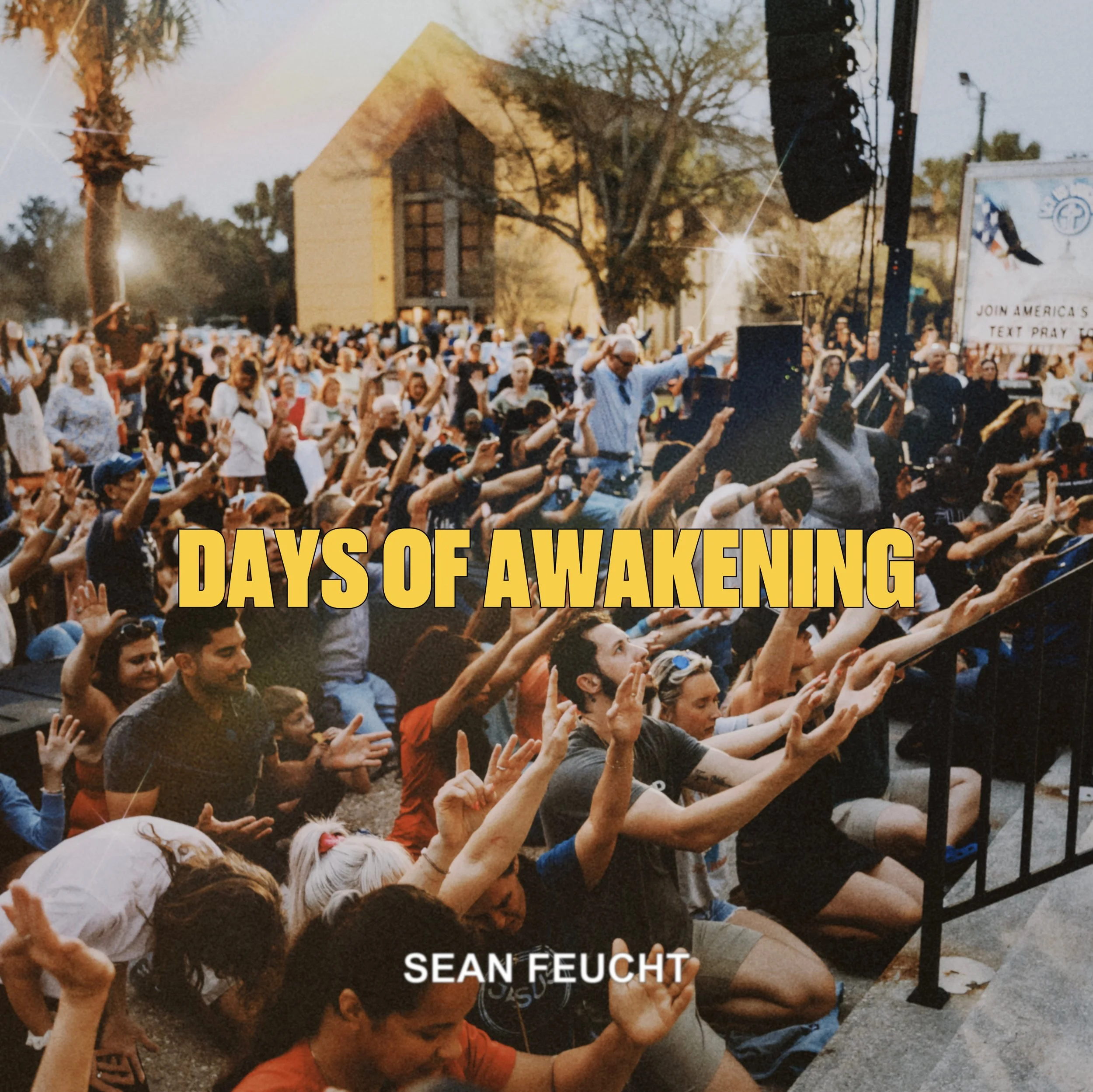 A large crowd of people gathered outdoors with many raising their hands, participating in an event called 'Days of Awakening' by Sean Feucht. The background features trees, a building, and stage equipment, with the scene captured during sunset or dus