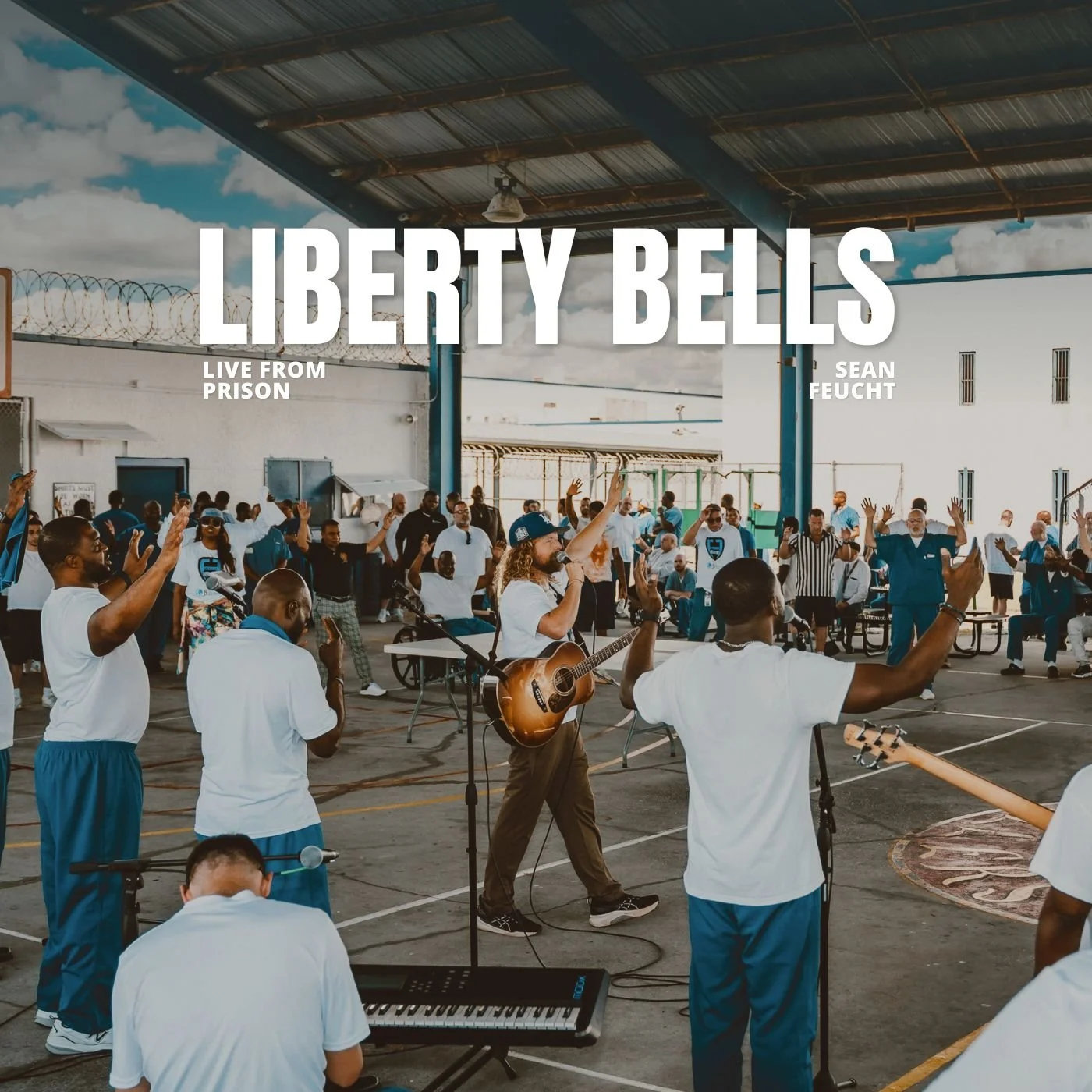 A group of people in prison gathered in an outdoor area with a partly cloudy sky, participating in a music event with individuals playing instruments and singing, some raising their hands, and a person in the center playing guitar.