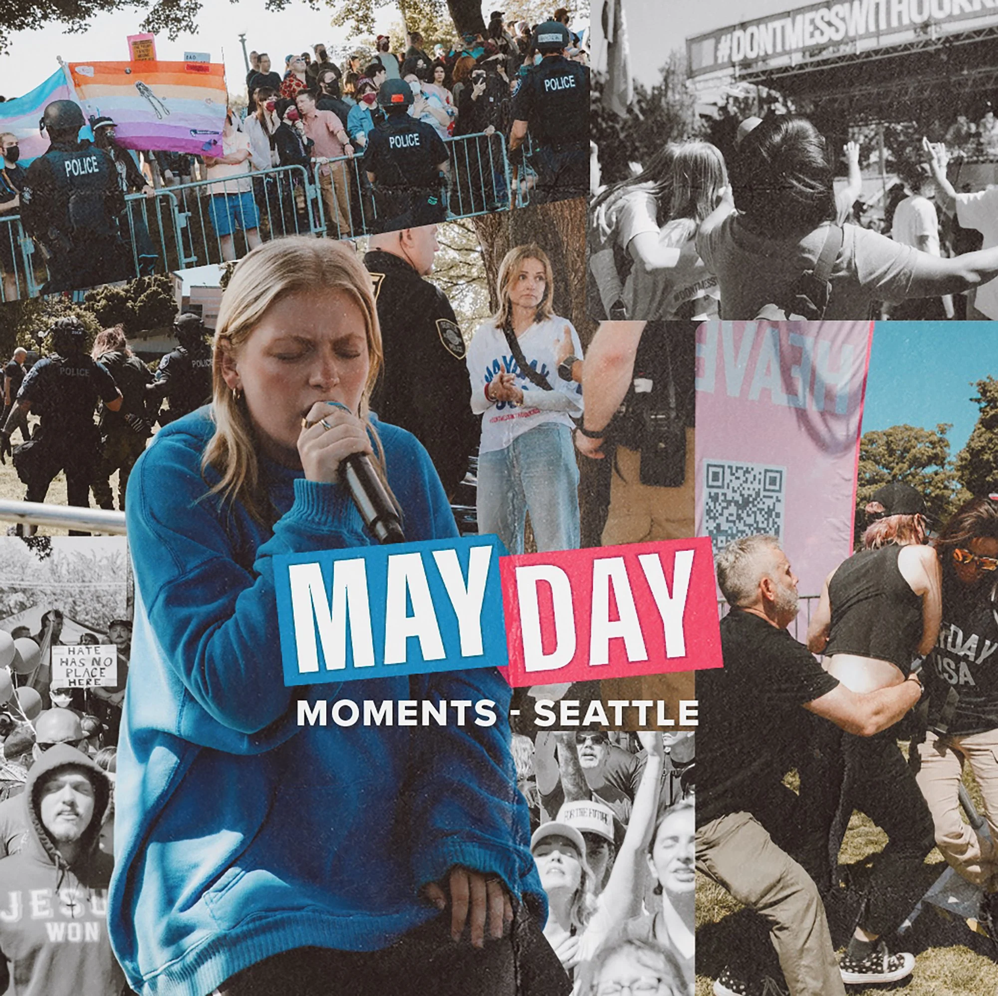 Woman in blue jacket speaking into a microphone at a May Day protest in Seattle with diverse crowd holding signs and flags, some with police presence.