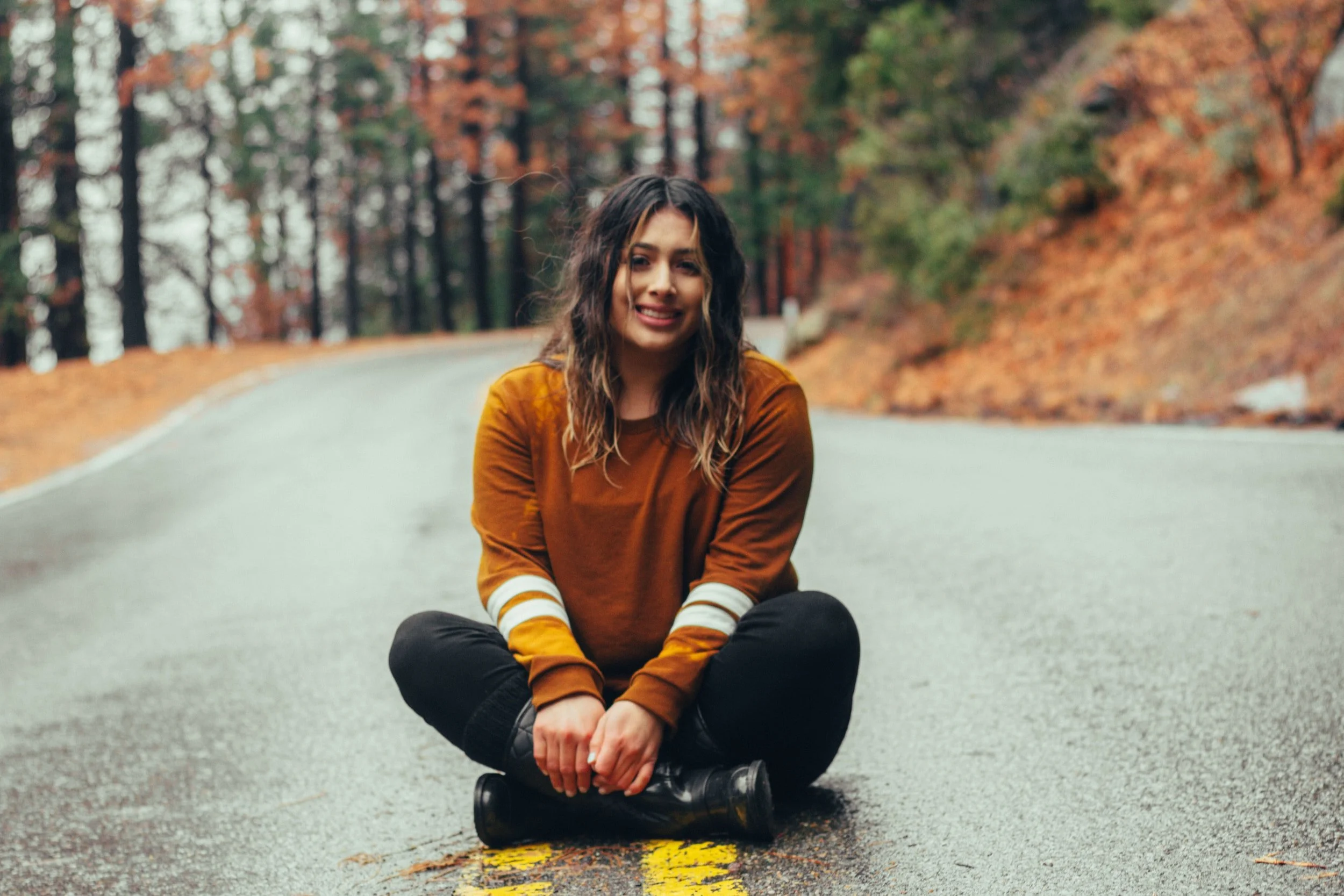 A young woman with wavy brown hair sitting cross-legged on a wet road in a forest with autumn foliage, smiling at the camera.