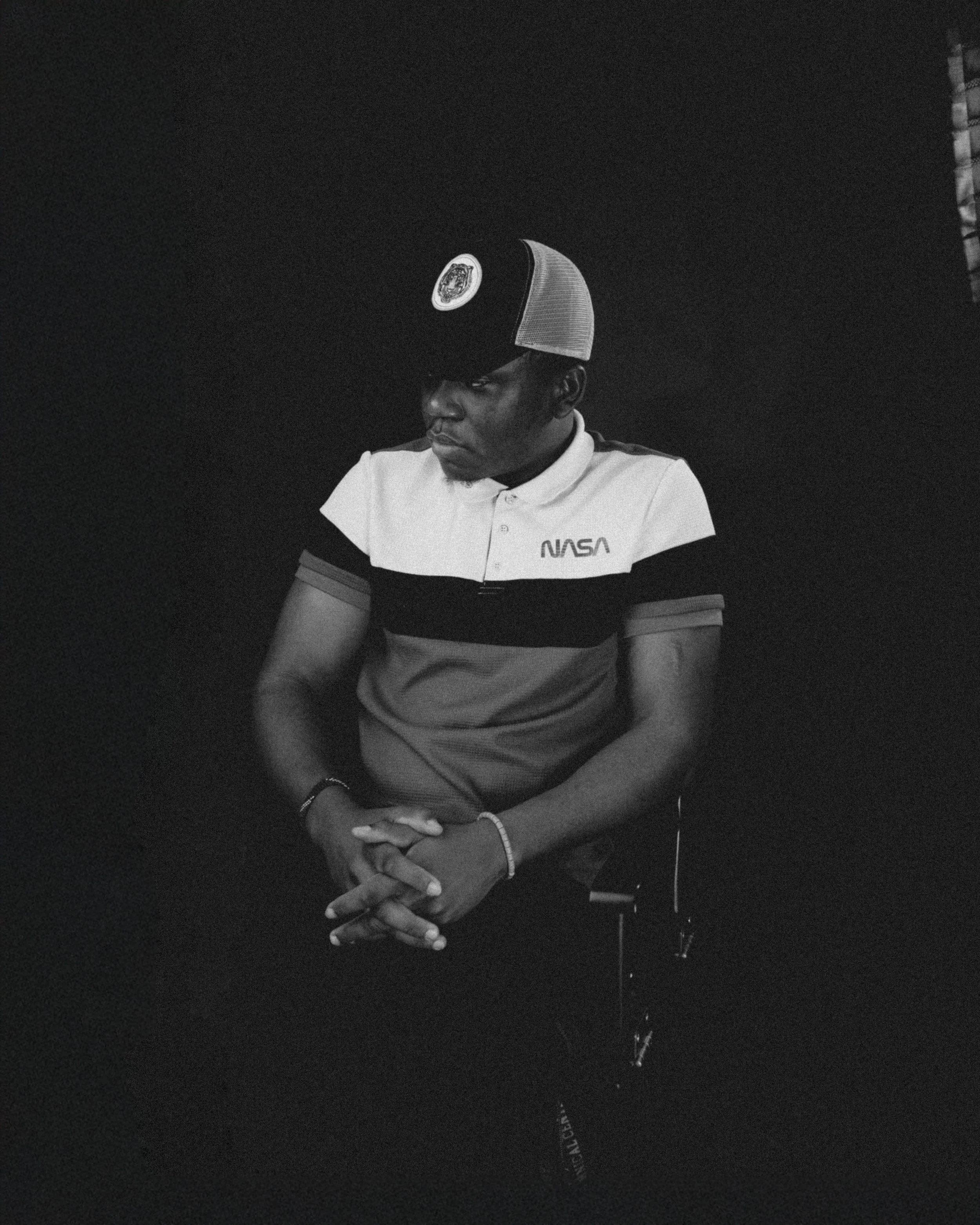 A young man wearing a NASA polo shirt and a baseball cap with a NASA emblem, sitting with his hands clasped, looking downward in a dark setting.