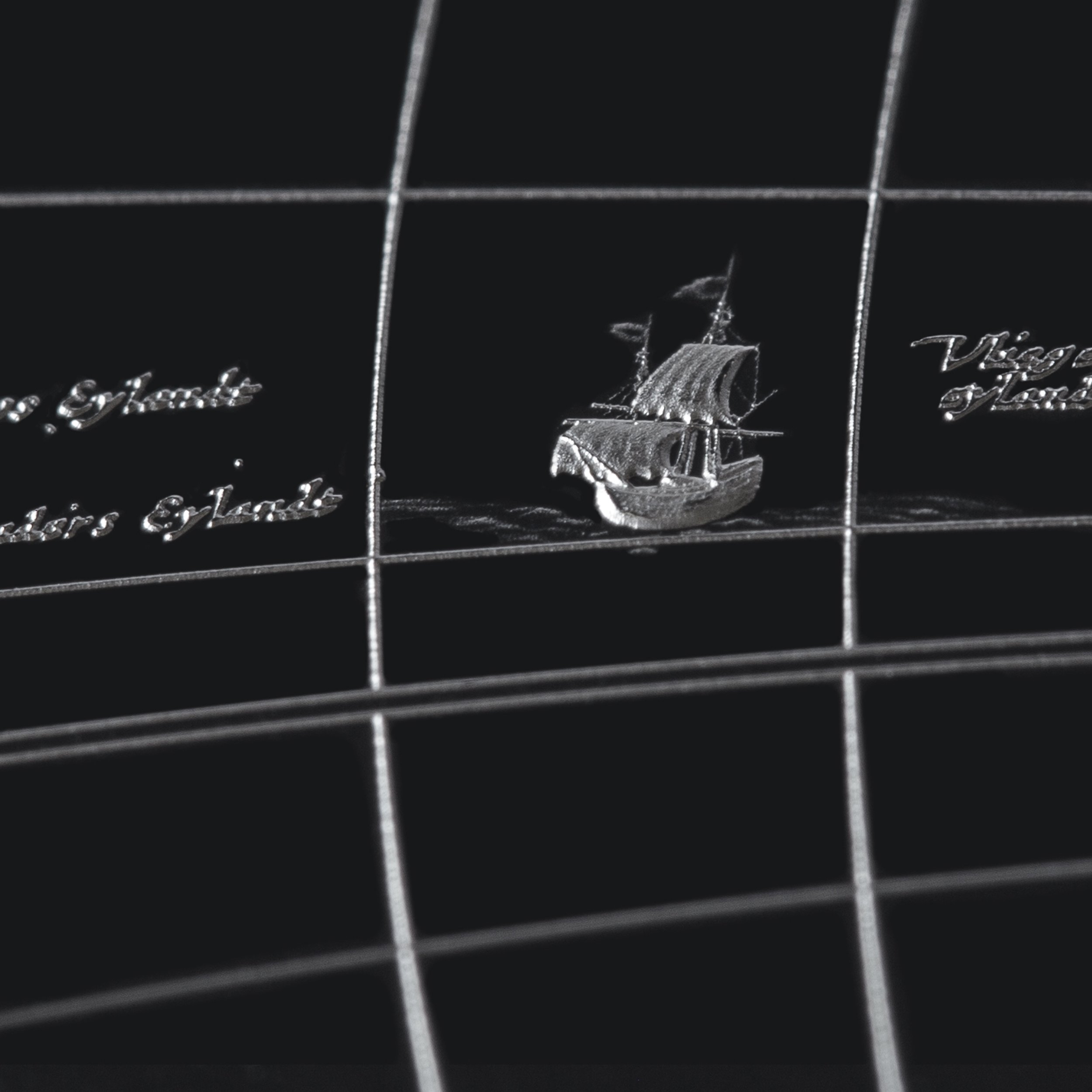 Close-up of a silver embossed sailing ship on a black surface divided by grid lines, with some cursive writing nearby.