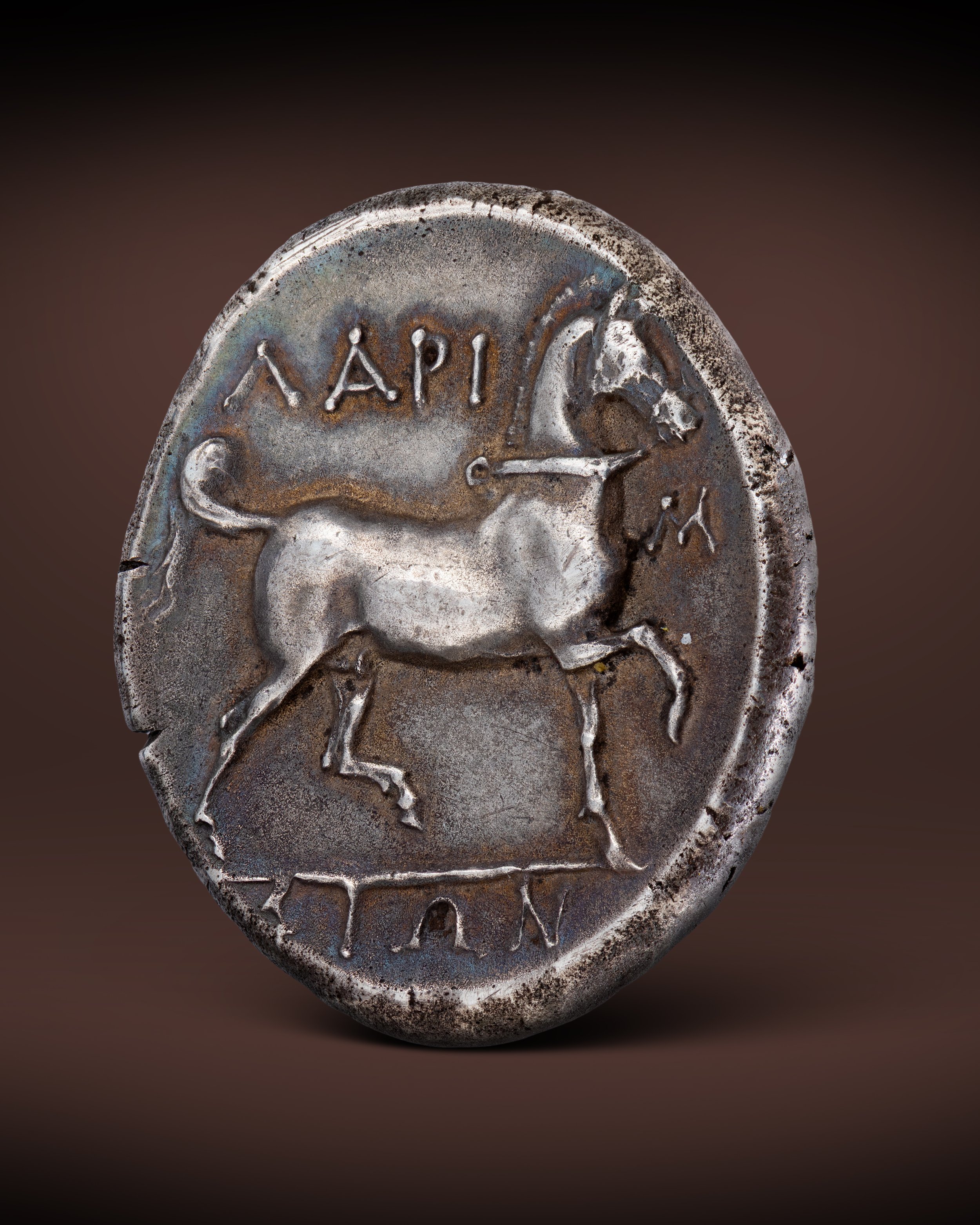 Ancient silver coin with Greek inscriptions and a profile profile of a female figure, possibly a goddess, with part of the inscription visible at the top and bottom.