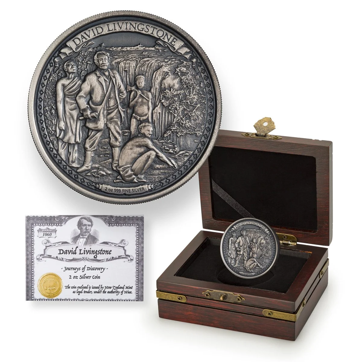 A silver coin honoring David Livingstone, featuring a detailed image of him with children in front of waterfalls, displayed in an open wooden box with an accompanying certificate of authenticity.