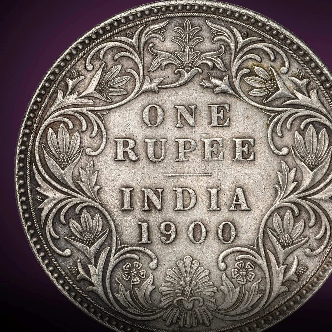 Close-up of an Indian one rupee coin from 1900, featuring intricate floral engravings around the text 'ONE RUPEE INDIA 1900'.