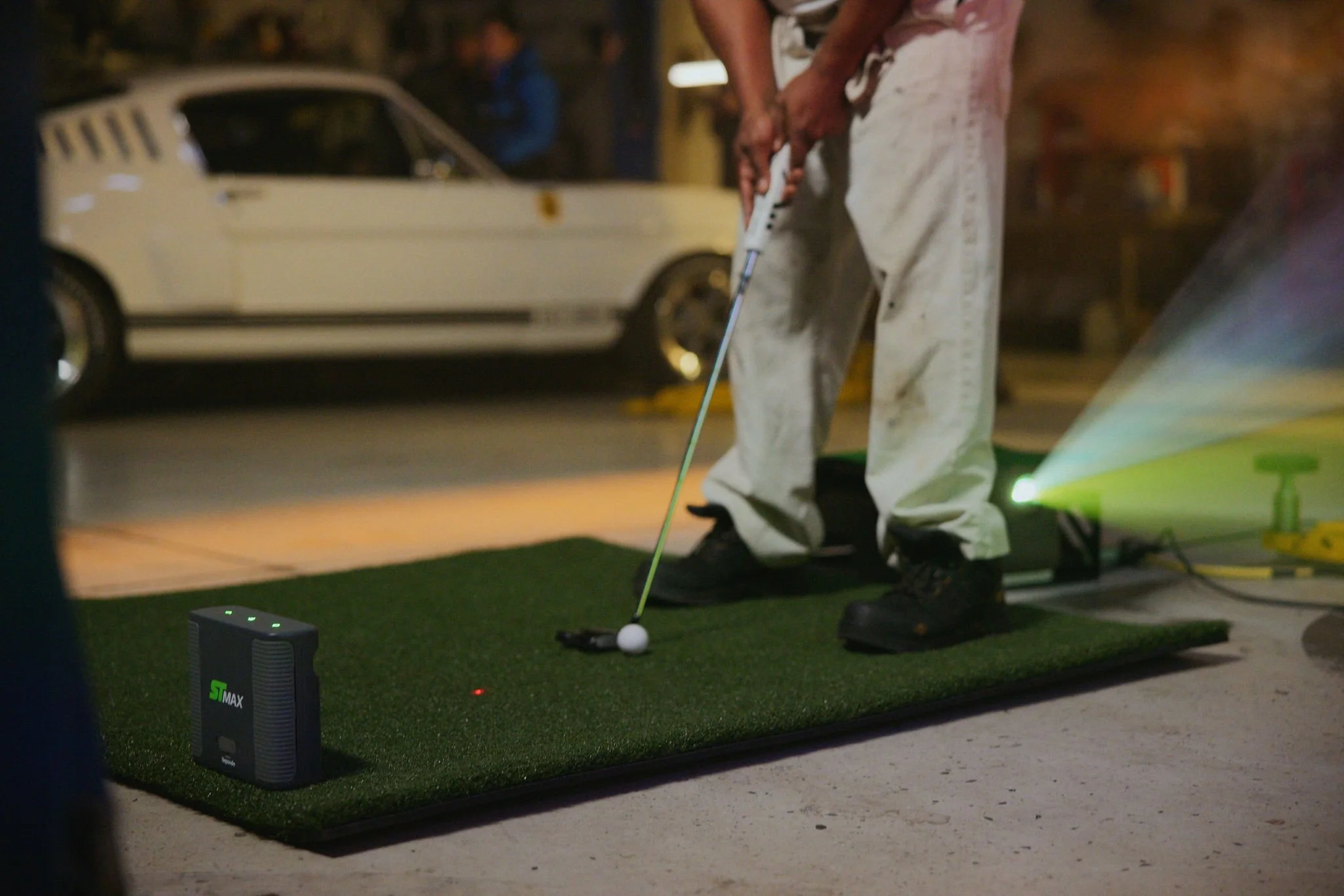 Person holding a golf club in a mechanic's garage setting, playing a golf simulator, with a white classic car in the background, and golf simulator equipment on the ground.