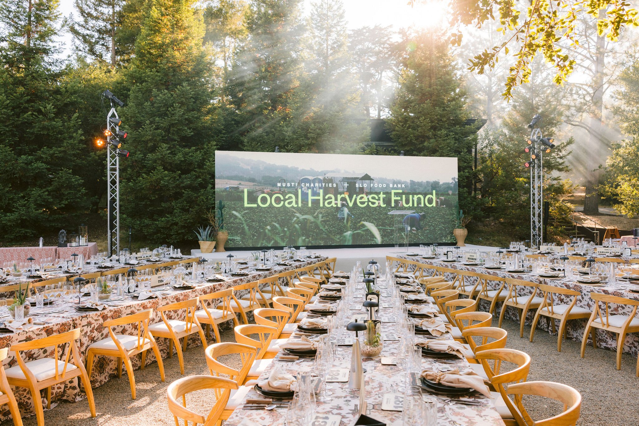 Outdoor event setup with long banquet tables decorated with floral tablecloths, surrounded by wooden chairs, with a large screen displaying "Local Harvest Fund" in front of a wooded area. Sunlight filters through the trees, creating a warm atmosphere