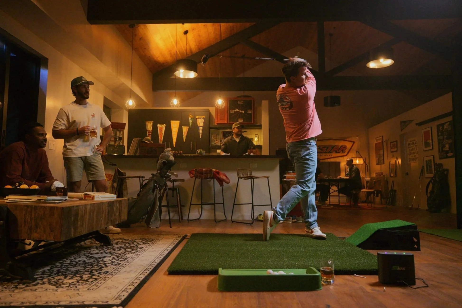 A man in a pink shirt swinging a golf club on an indoor putting green golf simulator, with several people seated and standing in a cozy, dimly lit room decorated with posters and warm lighting.