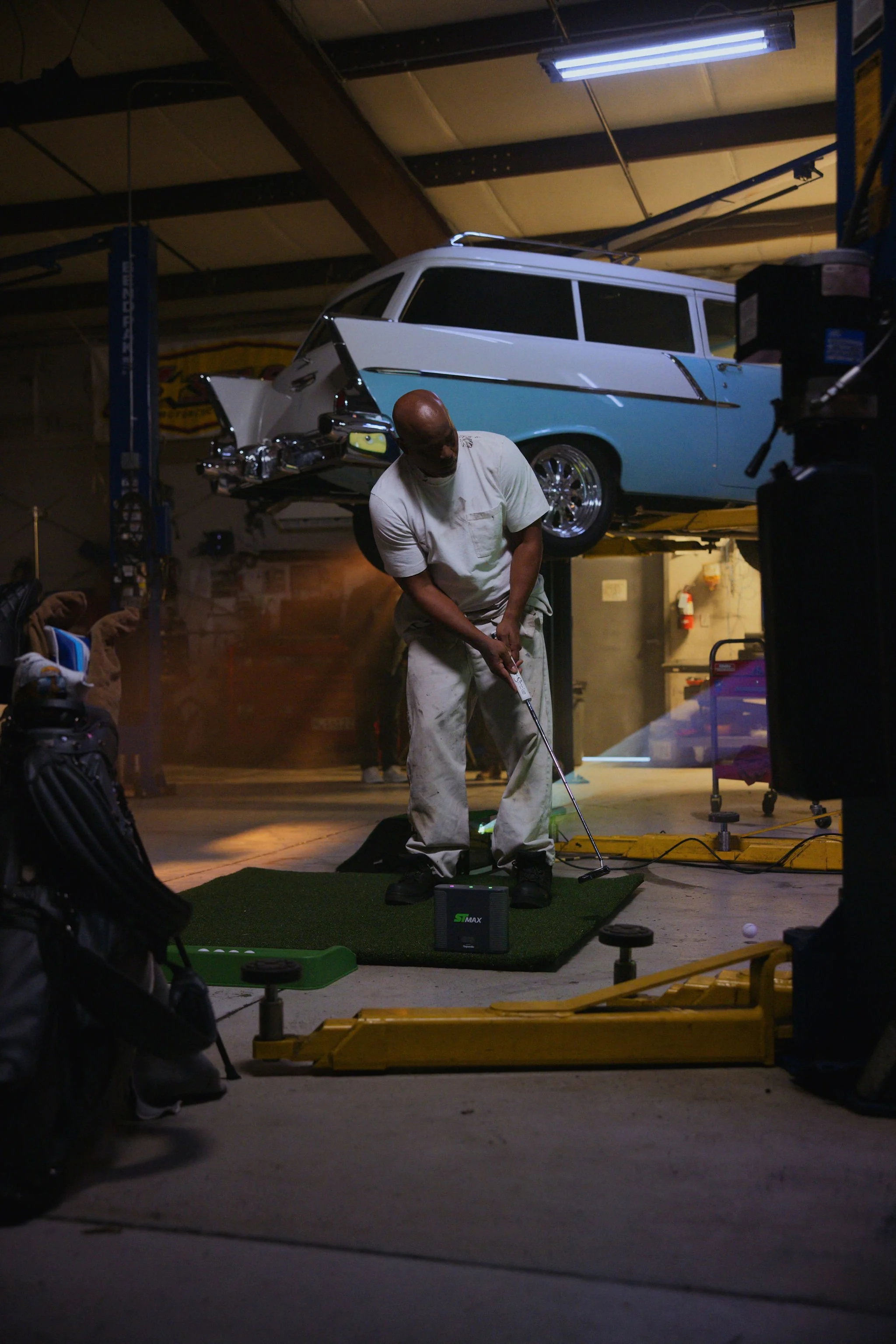 A mechanic man in white clothes practicing a golf simulator in his mechanic's shop, with a golf ball and a golf net visible, and a vintage station wagon on a lift in the background.