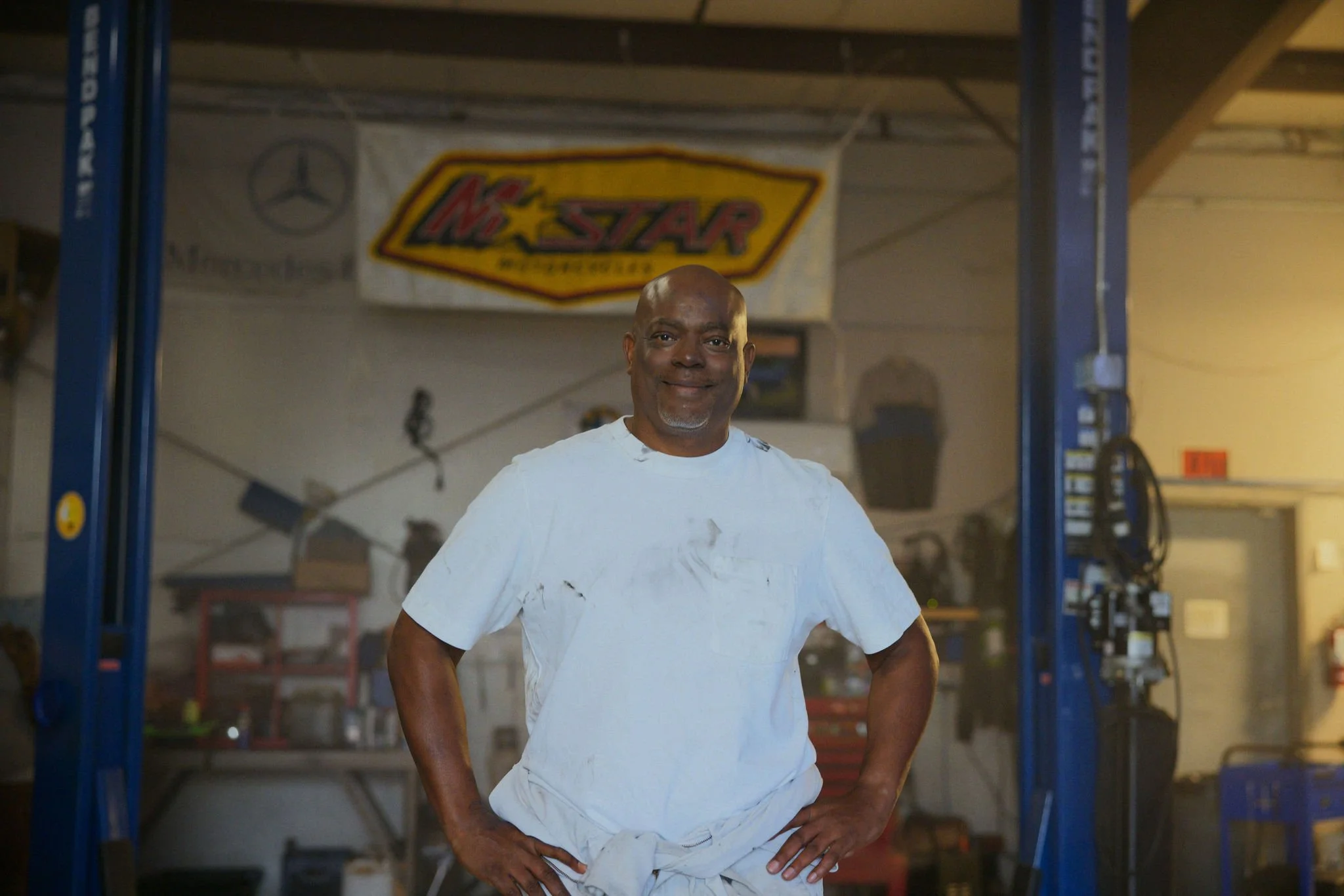 A smiling African American man mechanic with a shaved head and gray goatee, wearing a white t-shirt, standing in a garage or workshop with a motorcycle lift and shelves in the background.