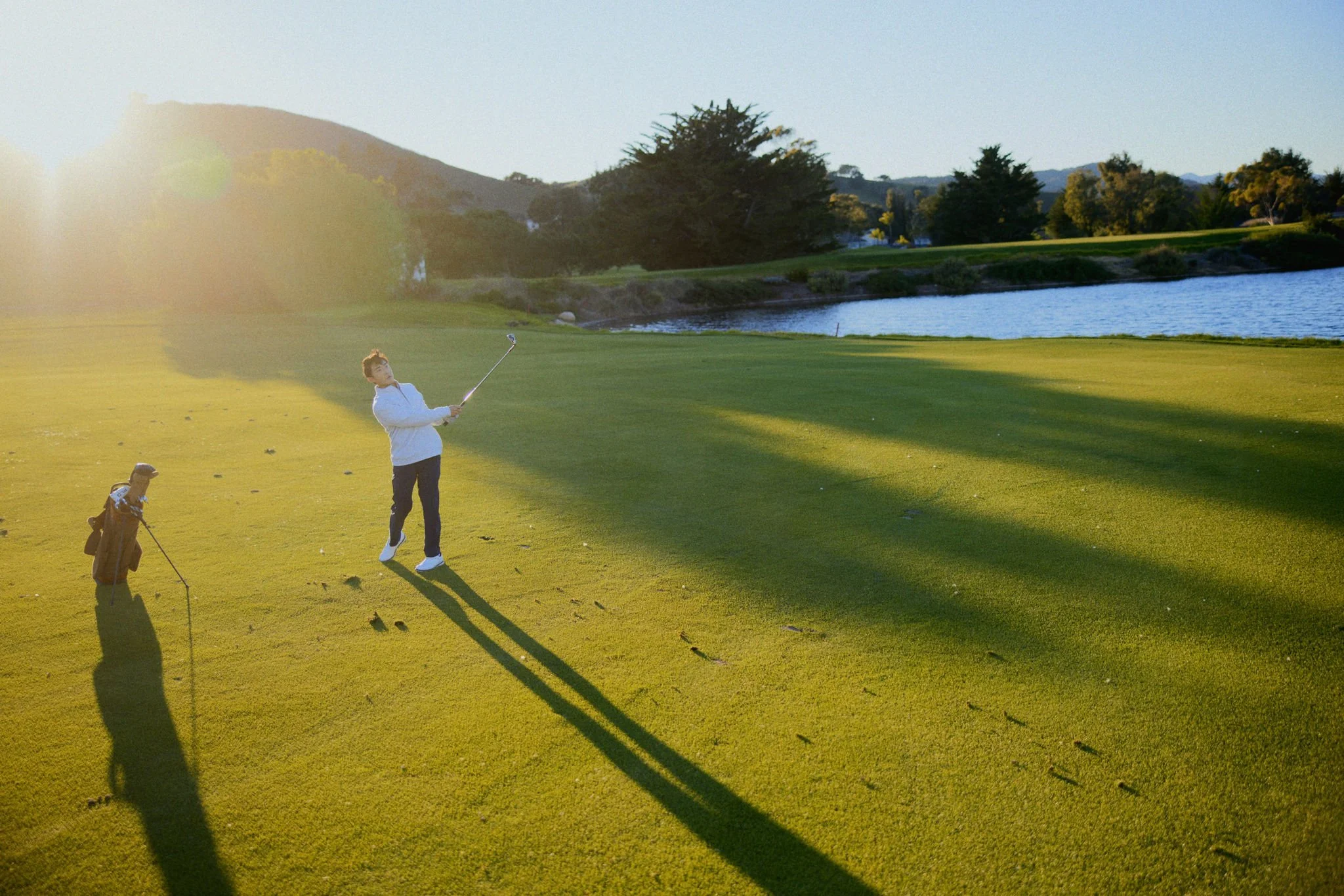 A man playing golf on a course near a body of water during sunset, with trees and hills in the background.
