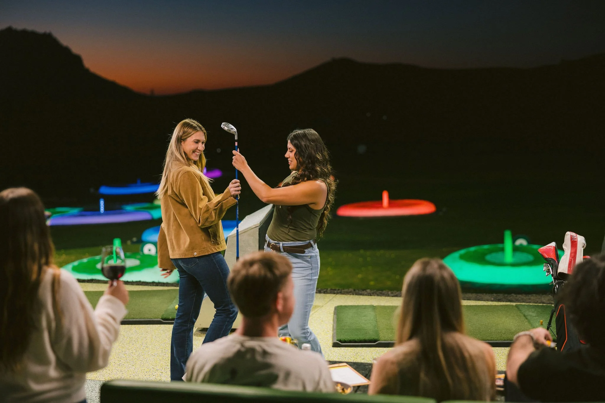 A woman in a brown jacket receiving a golf club from another woman in a green tank top on a golf course at night, with a group of people watching, holding a glass of red wine, and colorful illuminated targets in the background.
