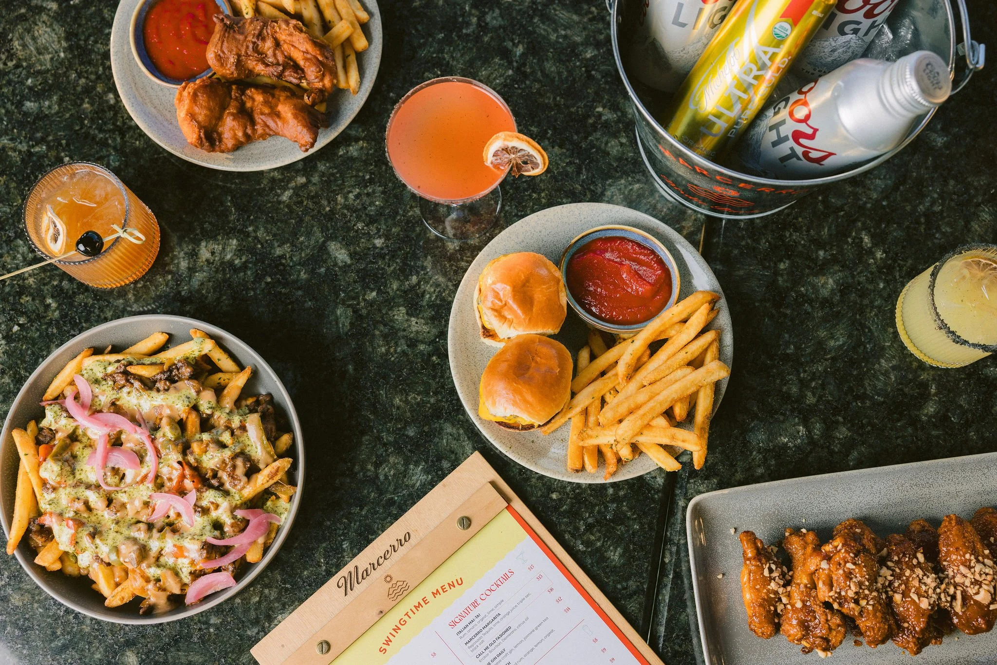 Marcerro Restaurant. Variety of dishes and drinks on a dark green marble table, including fried chicken, sliders with fries and ketchup, pasta with sauce and pickled onions, and assorted canned beverages at Swing Time in San Luis Obispo, CA