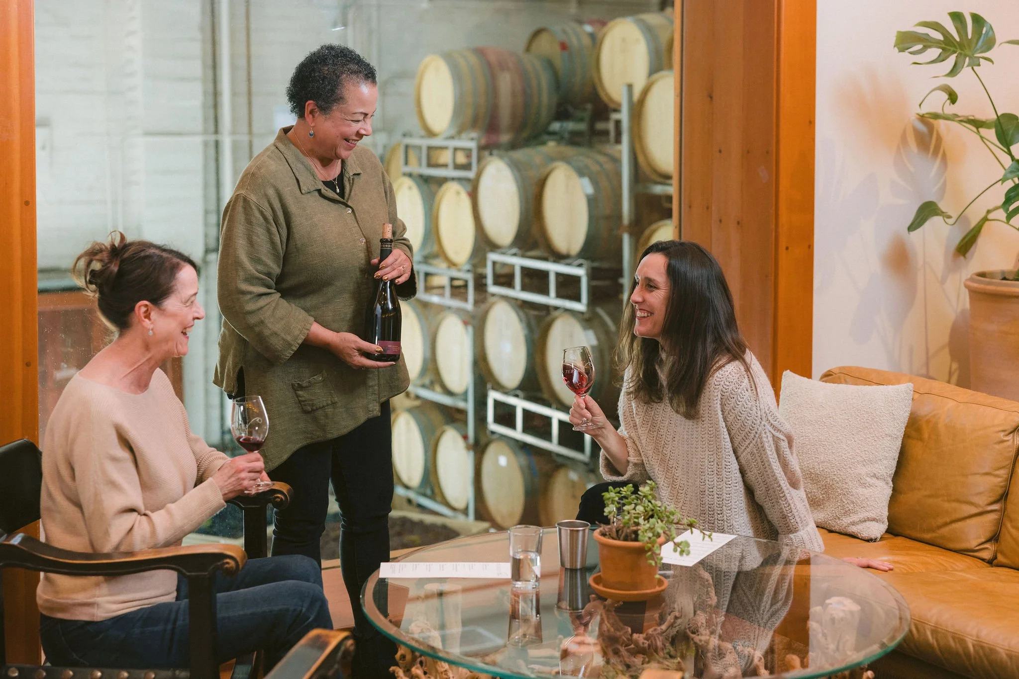 Three women in a winery seated around a glass table, having a wine tasting with a woman serving wine, with a window looking into a barrel room.