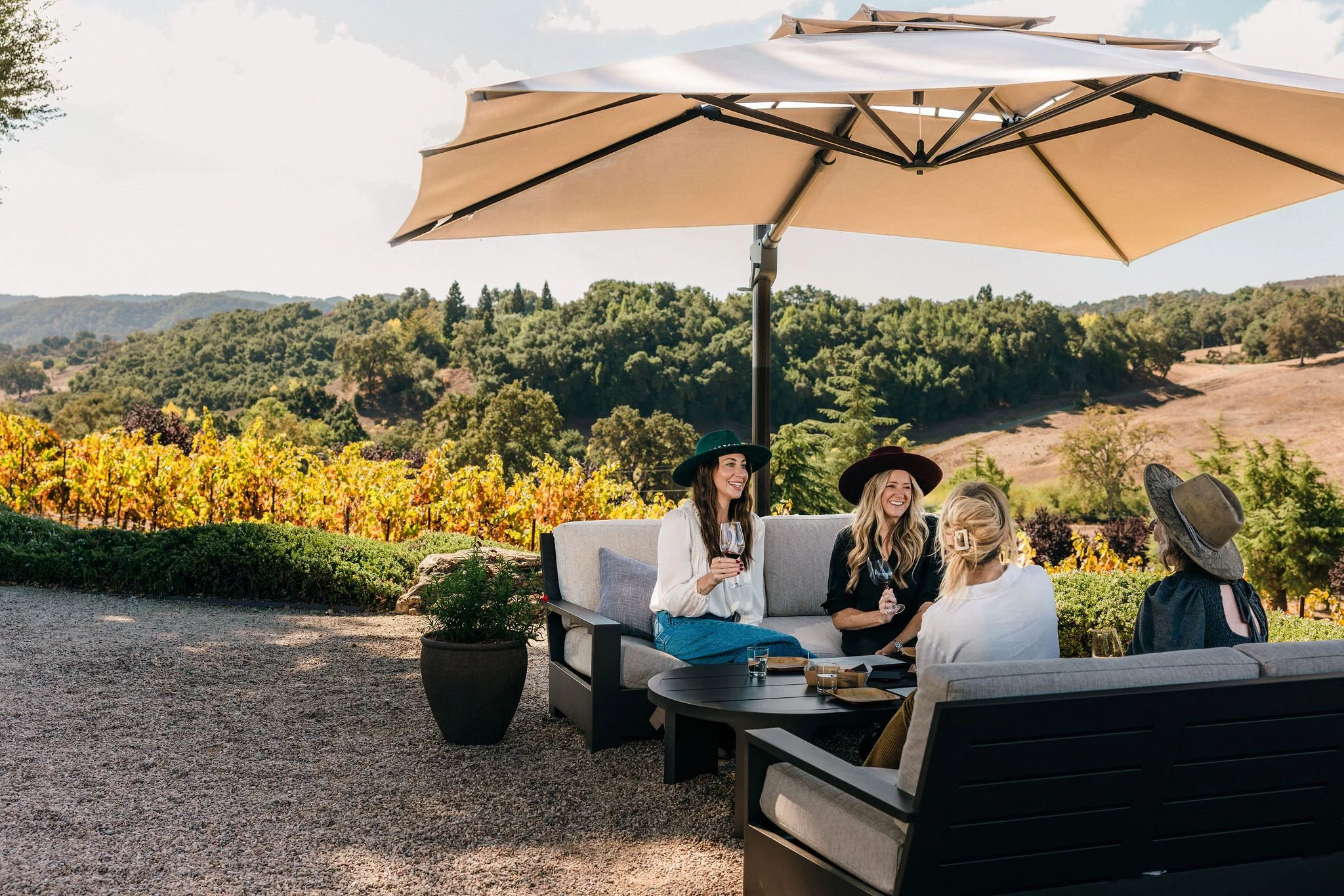 Four women sitting on outdoor patio furniture under a large beige umbrella, enjoying wine and conversation with a vineyard and hilly landscape in the background.