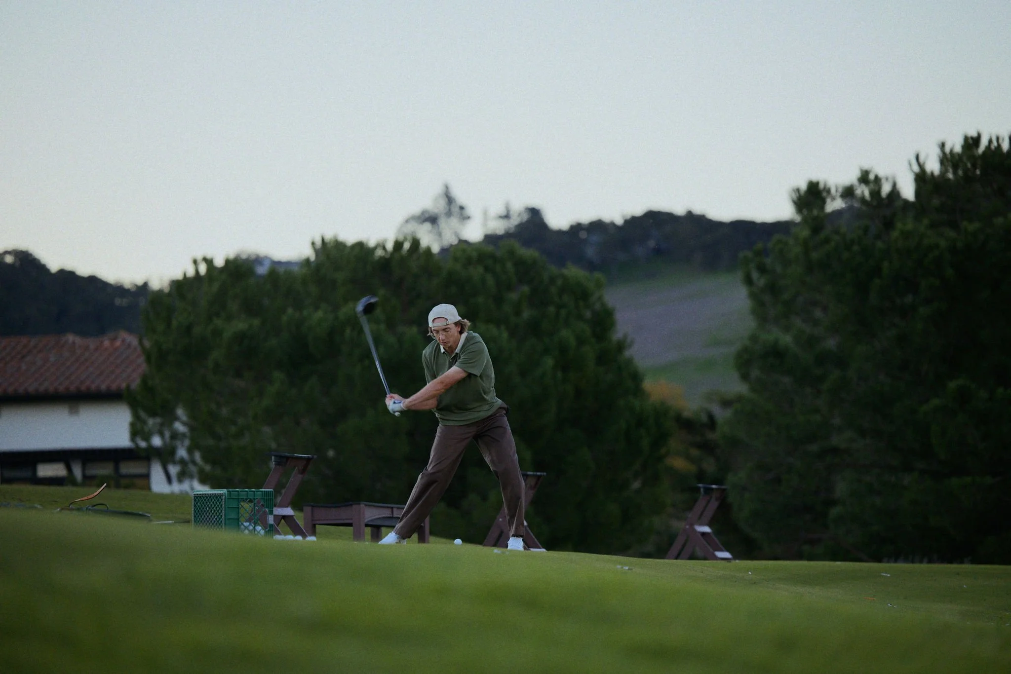 A man playing golf on a lush green country club course during the evening, preparing to hit a golf ball with a club, with trees and buildings in the background.