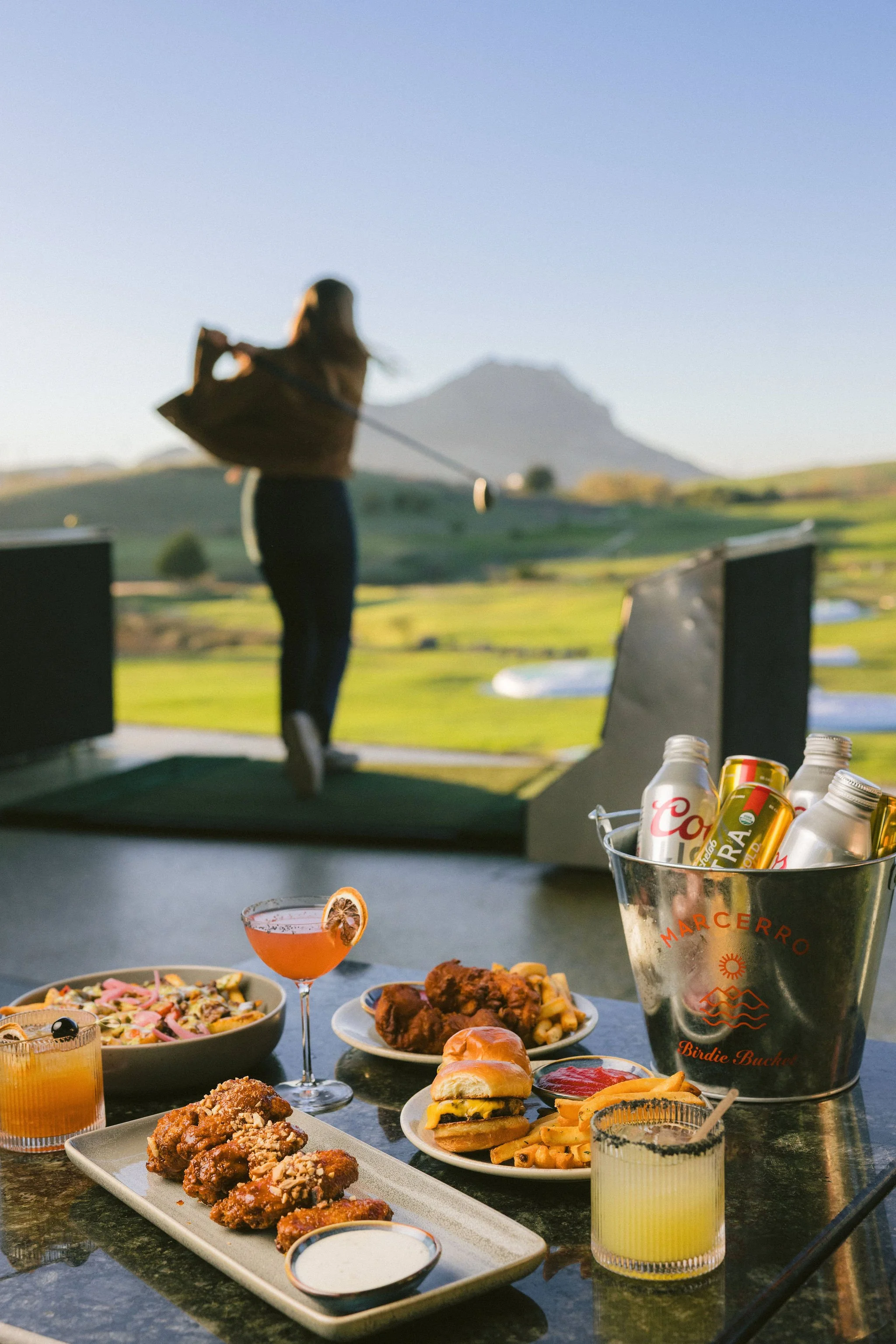 A woman playing top golf at Swing Time in San Luis Obispo with a spread of food and drinks on a table in the foreground, including fried chicken, burgers, fries, salads, cocktails, and beer.