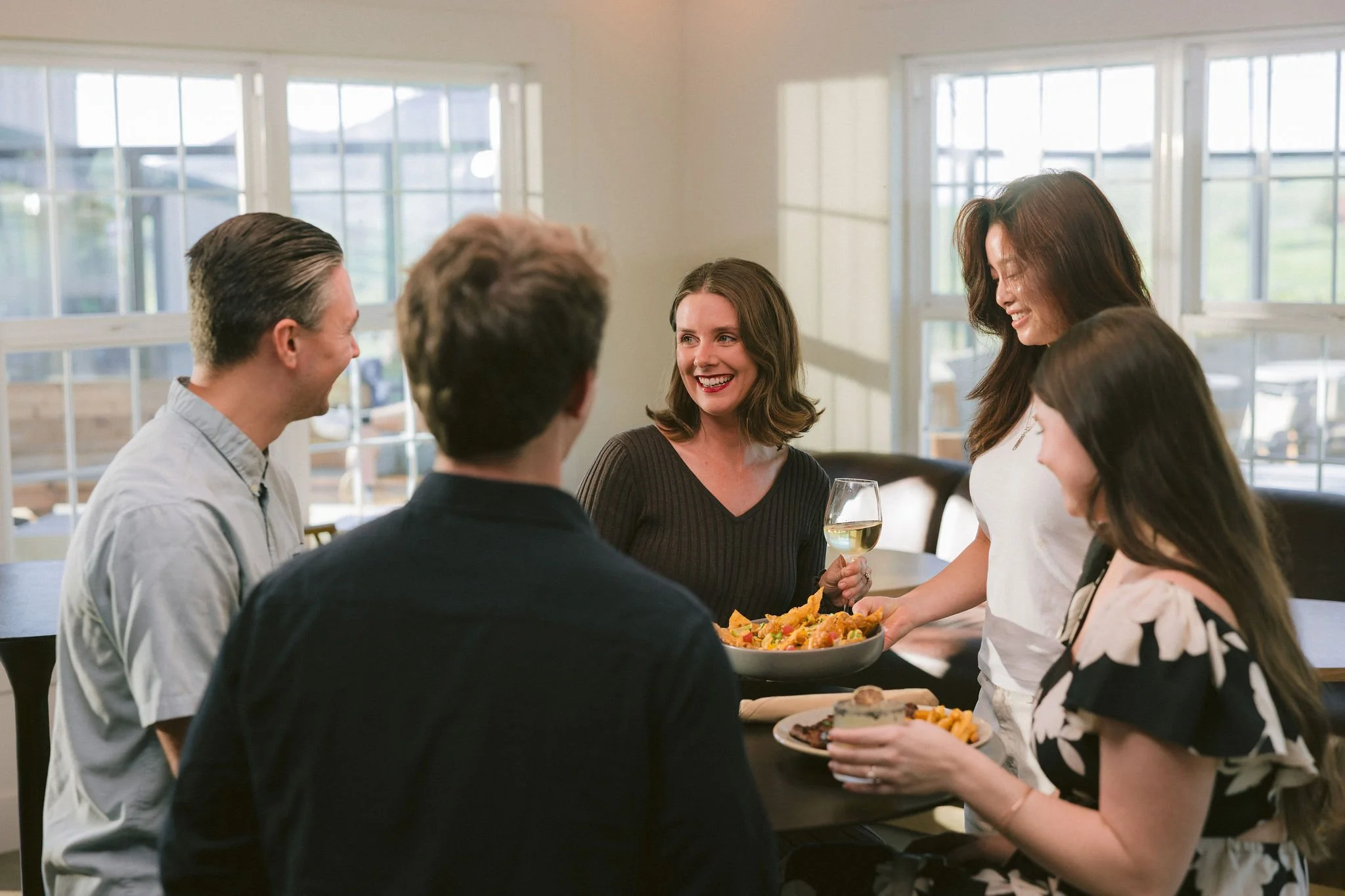 Group of five friends at a table enjoying a social gathering with food and drinks, smiling and chatting indoors near large windows.