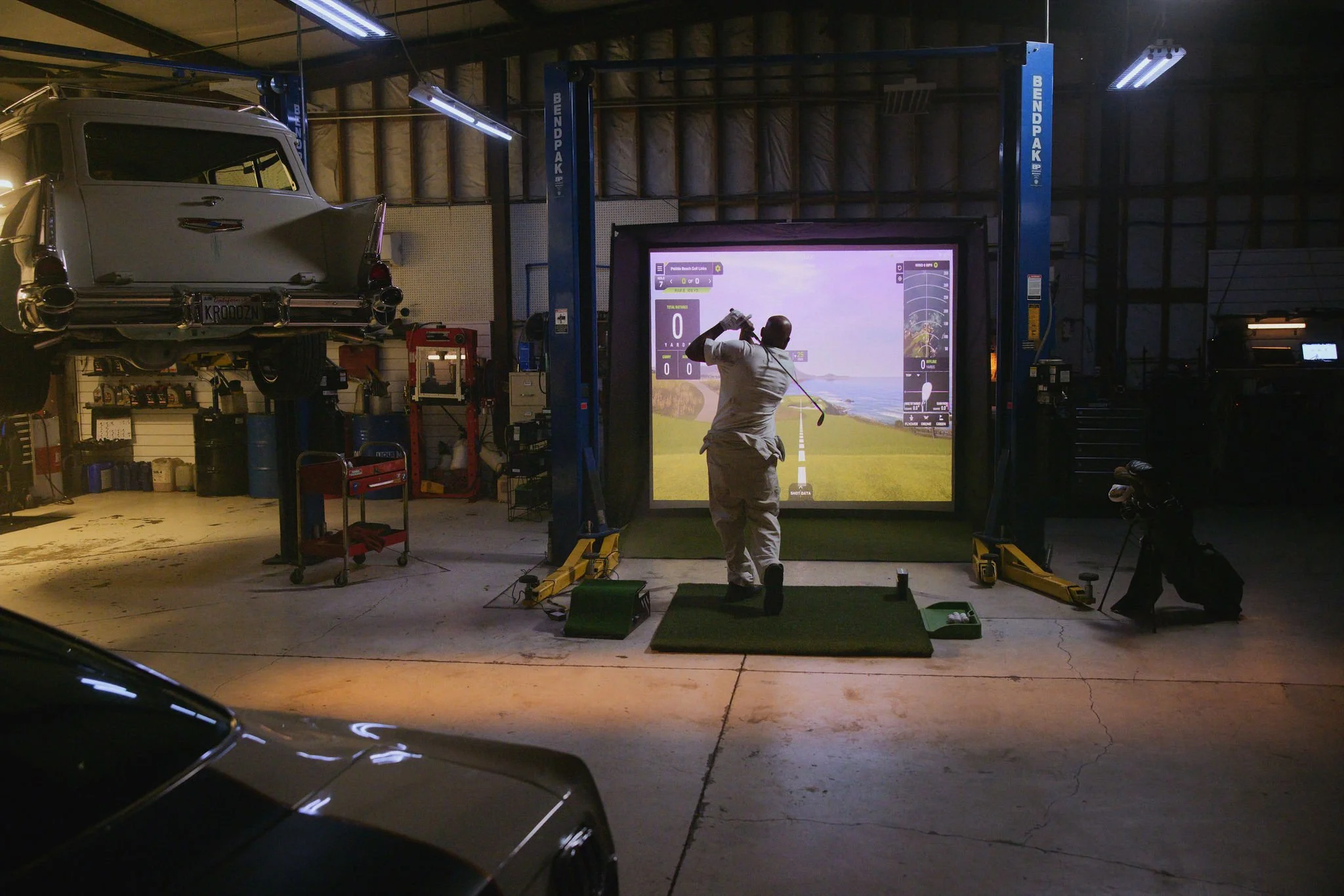A man practicing golf indoors on a simulator, standing on a mat with a golf club in his hands, facing a large screen displaying a scenic golf course and data.
