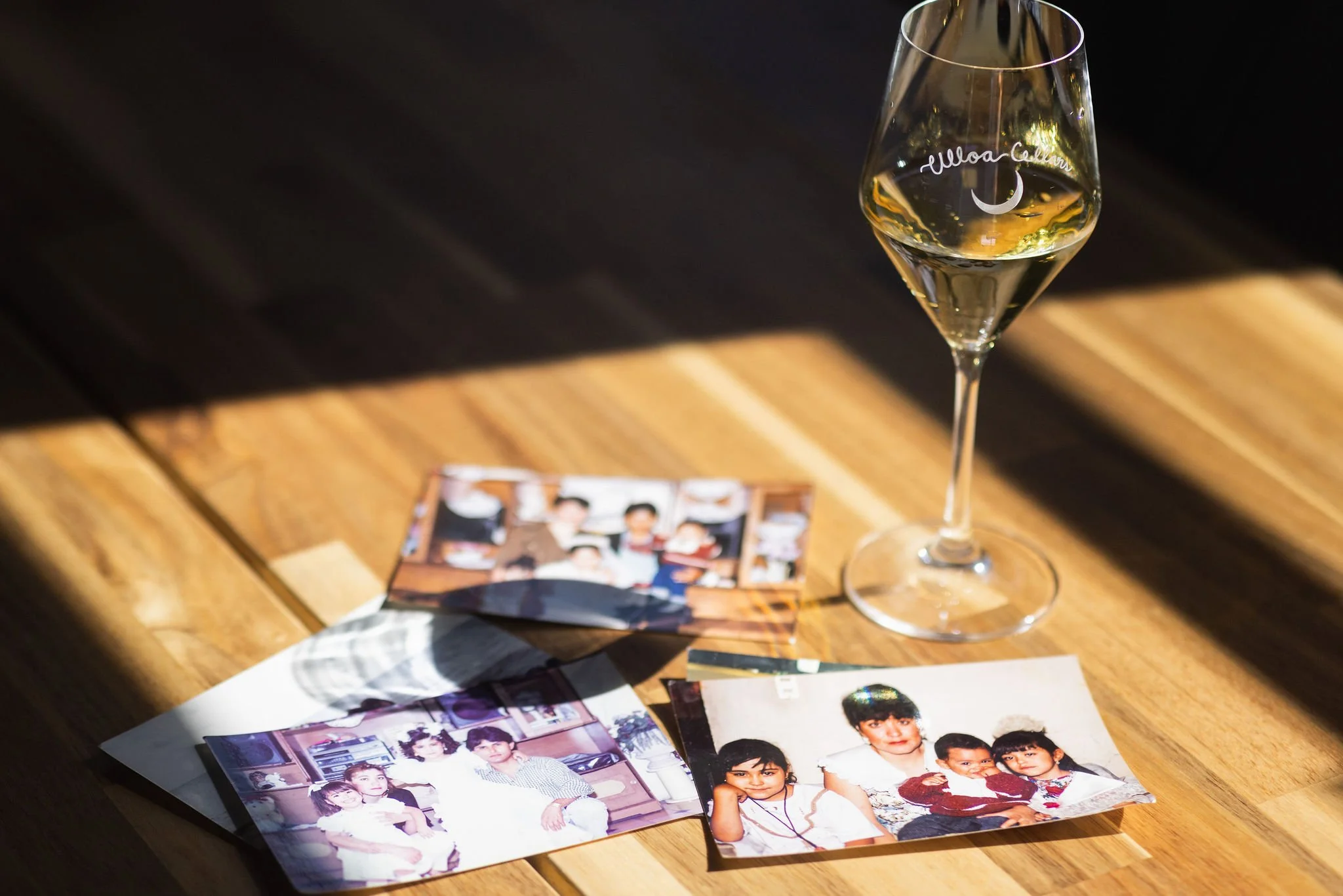 A stemmed glass with white wine on a wooden table, surrounded by several vintage family photographs.