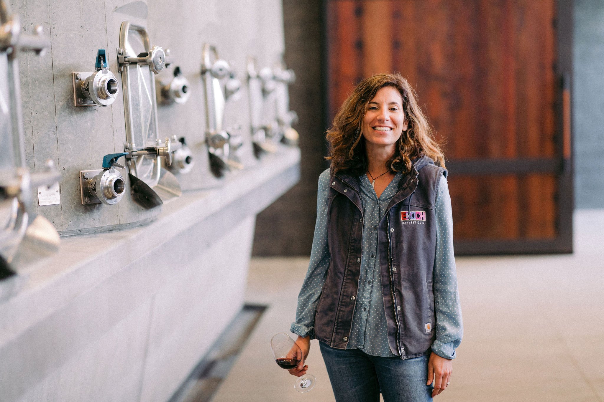 Smiling person holding a glass of red wine standing near industrial wine-making equipment.